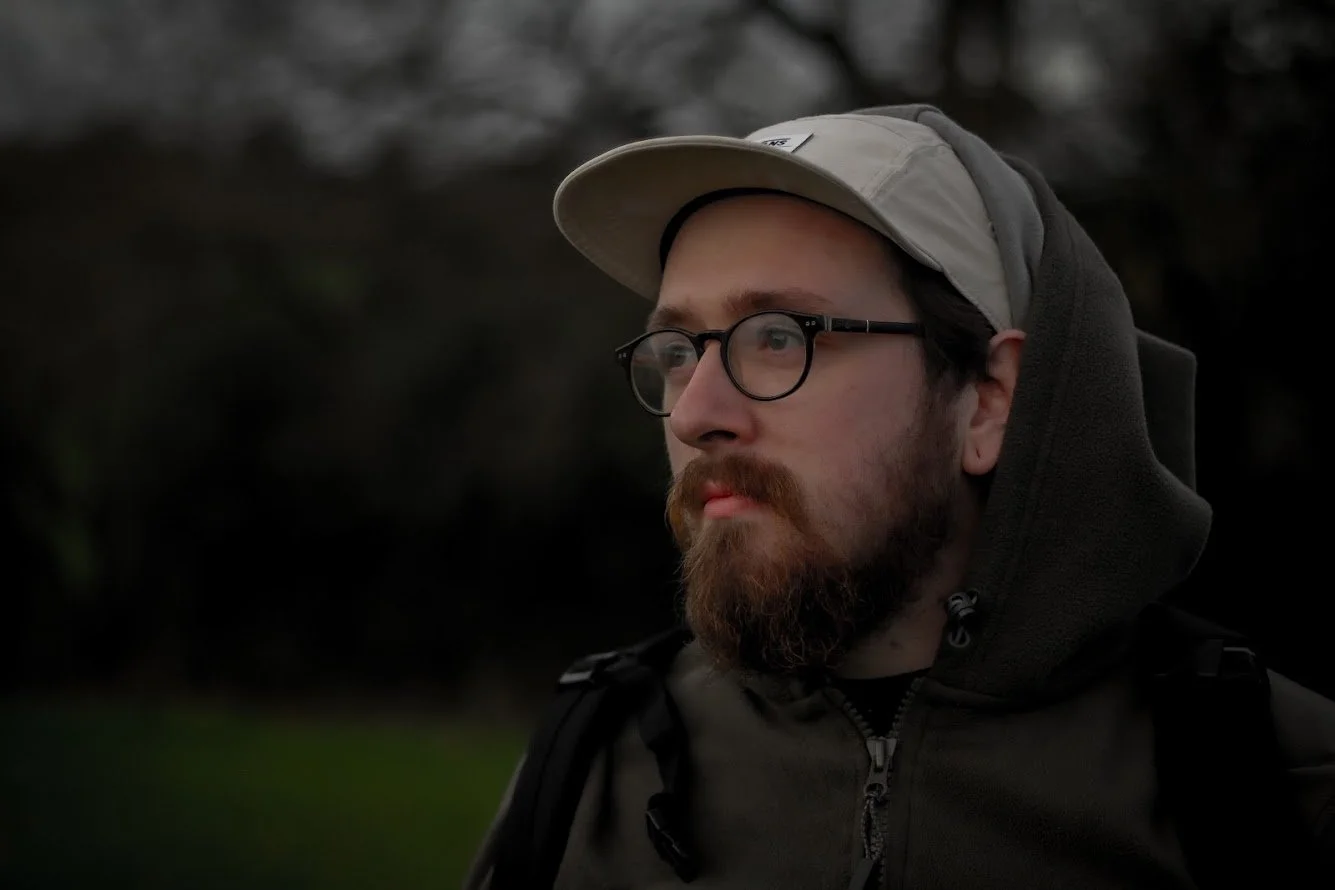 A man with glasses, a beard, and a mustache wearing a beige cap and a hooded jacket outdoors.