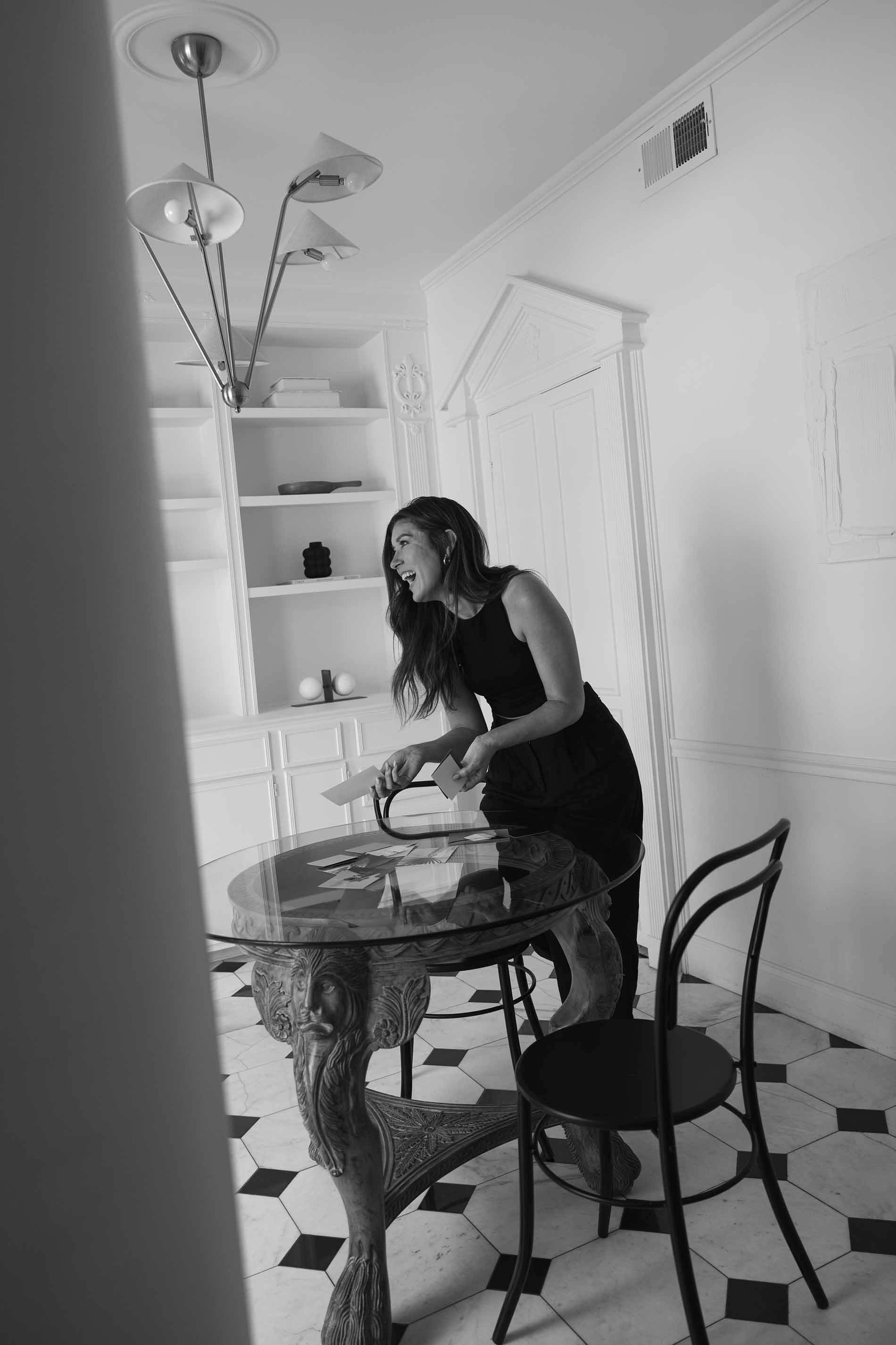 A woman in a black top and pants laughing while looking at photographs or postcards on a glass table in a dining room.