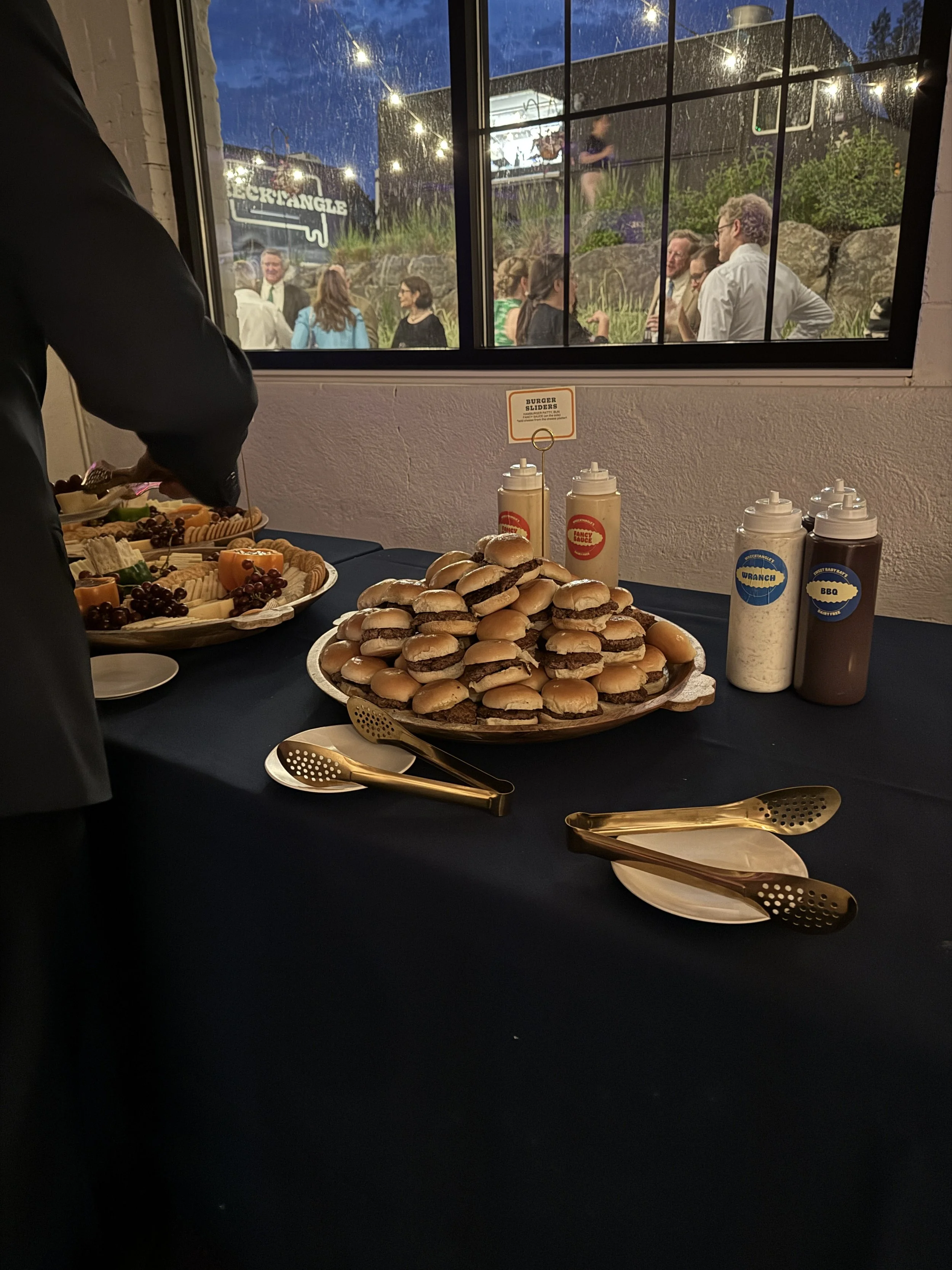 A table with mini cheeseburgers and a cheese platter with grapes and crackers, set for a social gathering, with condiments and plates arranged on a dark tablecloth.