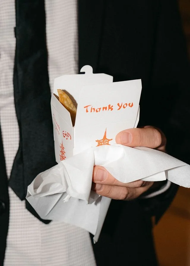 Person holding a French fry container with a thank you note, wrapped in a white napkin, person wearing a black jacket and vest.