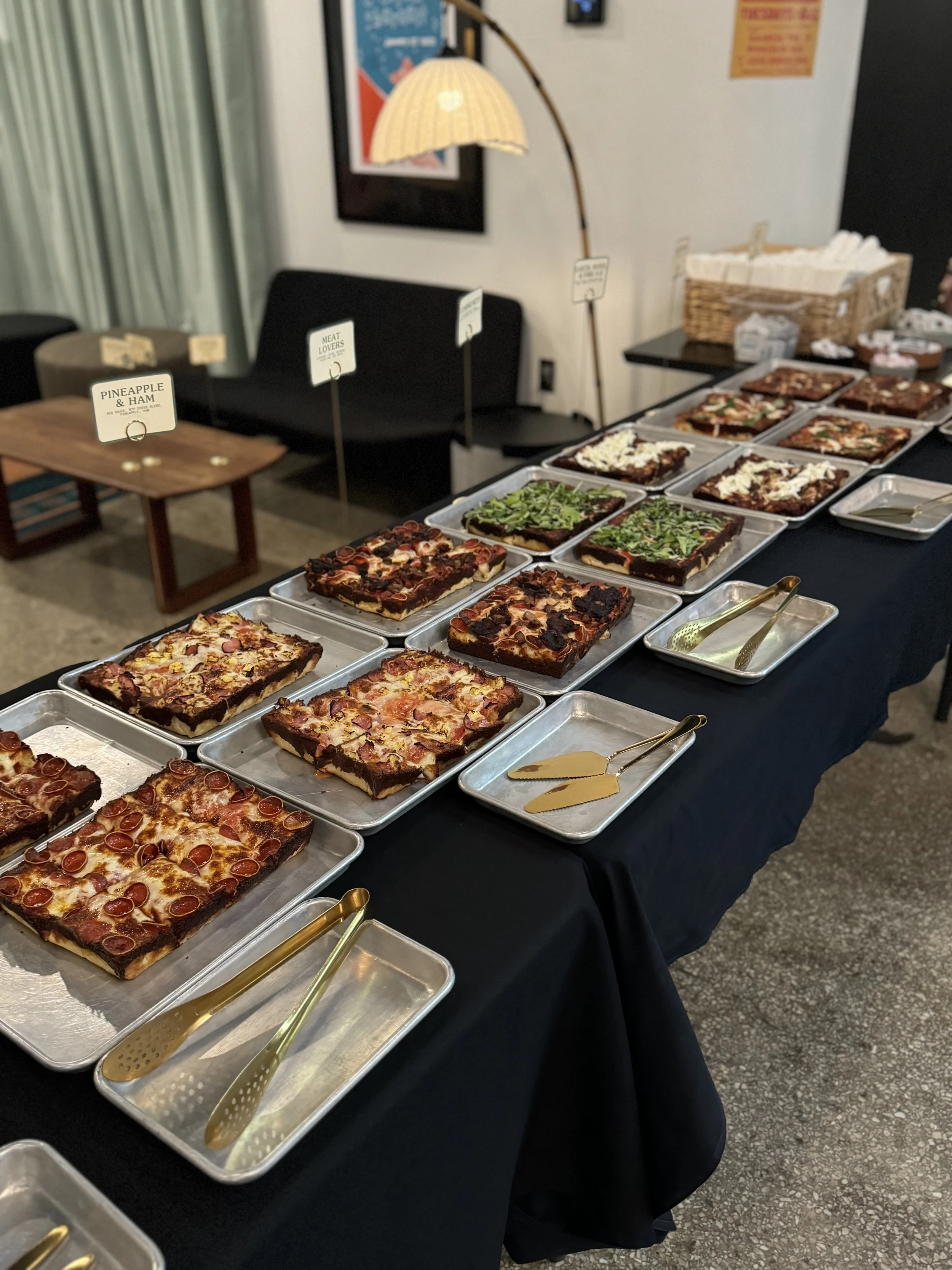 A table covered with various types of pizza slices on metal trays, with tongs in some trays, set up for a buffet or catering event.