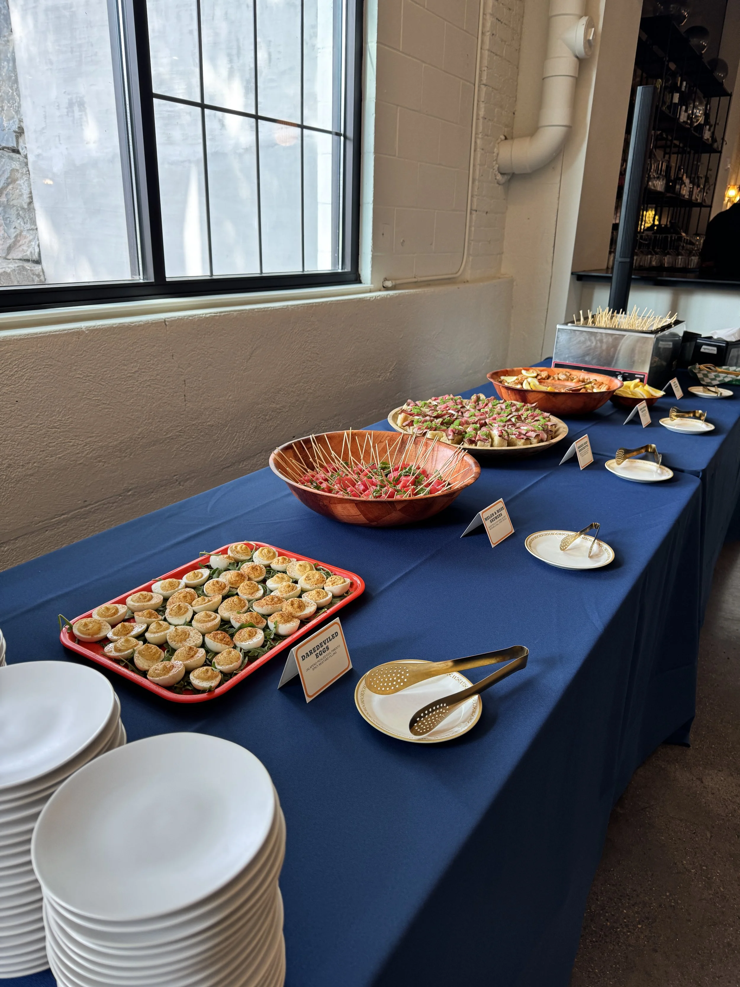 A buffet table with a navy blue tablecloth displaying various appetizers, including deviled eggs, skewers of meat and vegetables, and sliced fruit bowls, set next to a window with a white wall and gray piping.