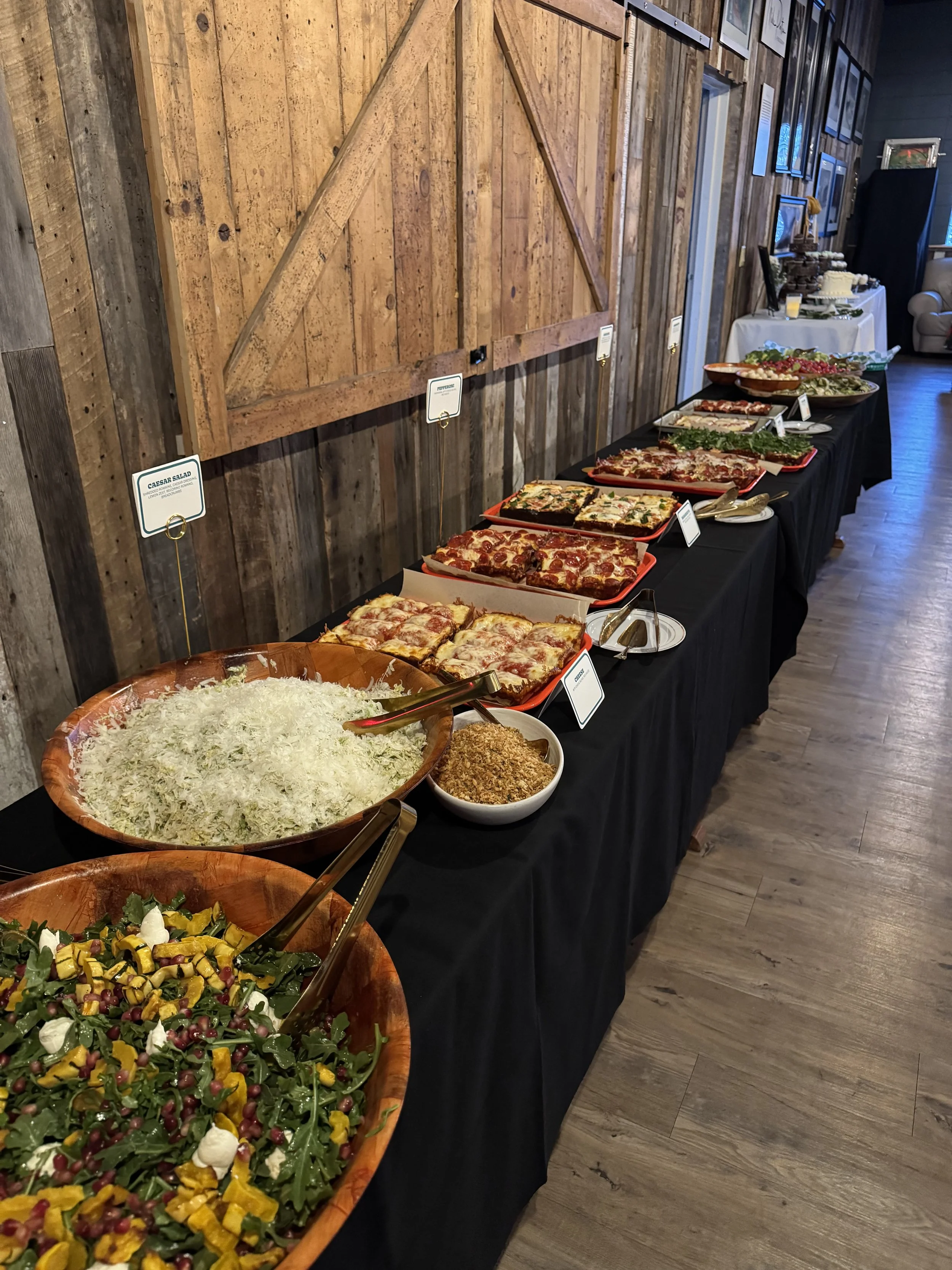 Buffet table with various salads, pizza, and appetizers, set against a rustic wooden wall, in an indoor setting.