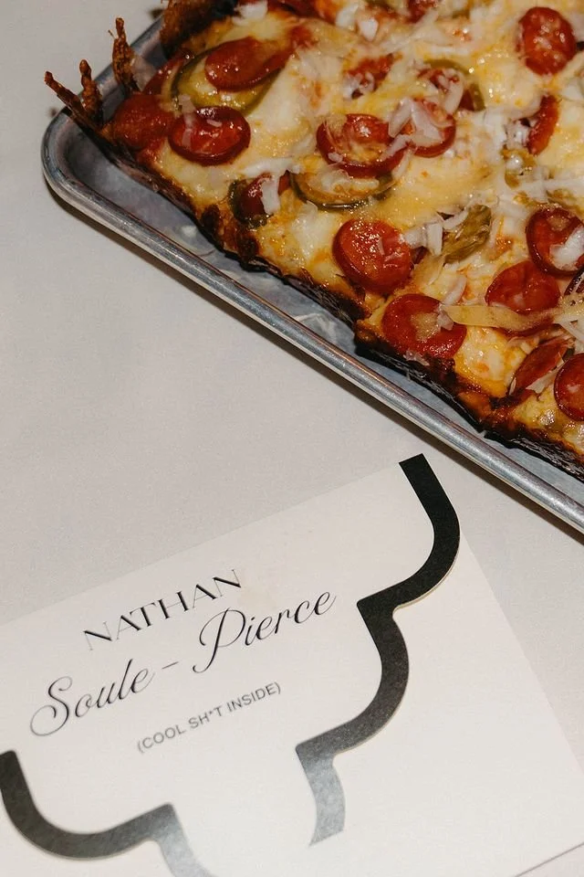 Close-up of a rectangular pepperoni pizza on a metal tray, with melted cheese, sliced onions, and green peppers. Part of a card with the name 'Nathan Soule-Pierce' and the note '(Cool sh*t inside)' is visible beneath the tray.