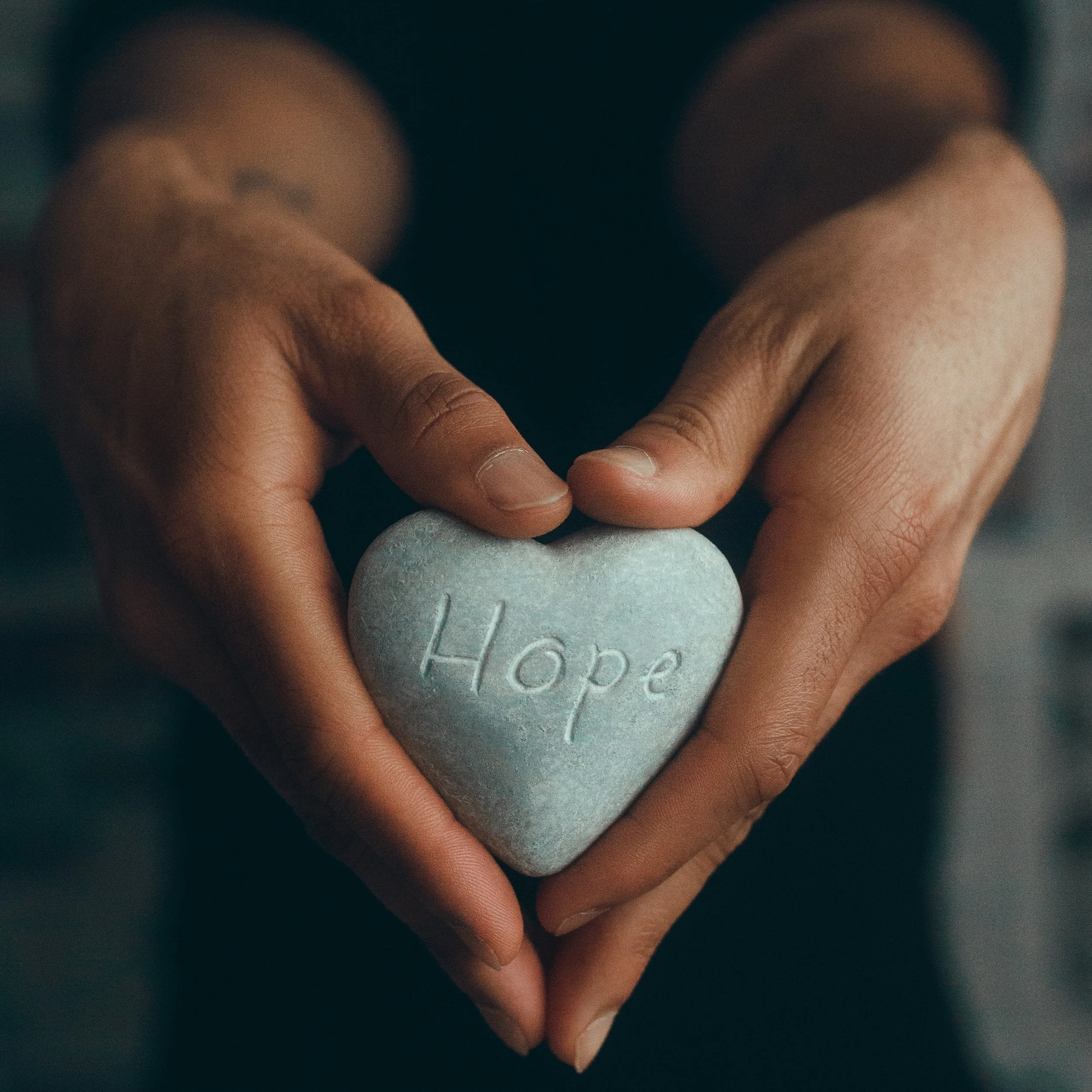 Hands holding a heart-shaped stone engraved with the word "Hope," symbolizing healing, compassion, and support.
