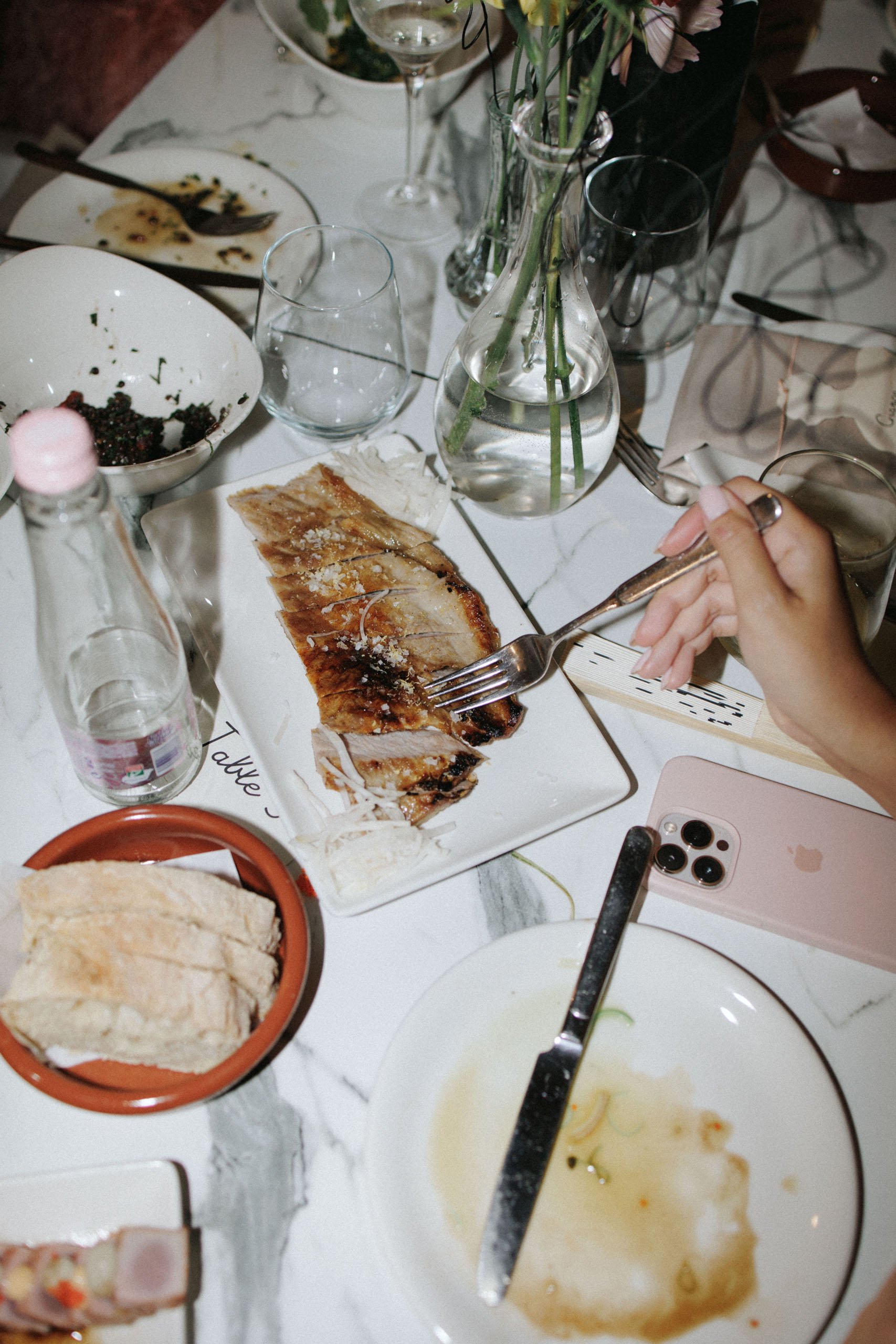 A nearly empty table with leftover food, including a plate of sliced roasted meat, a small terracotta dish with bread, a bowl with berries, and a few glasses and utensils, with a hand holding a fork. There is a pink iPhone, a glass jar with flowers, and other dishes and empty glasses scattered on the table.