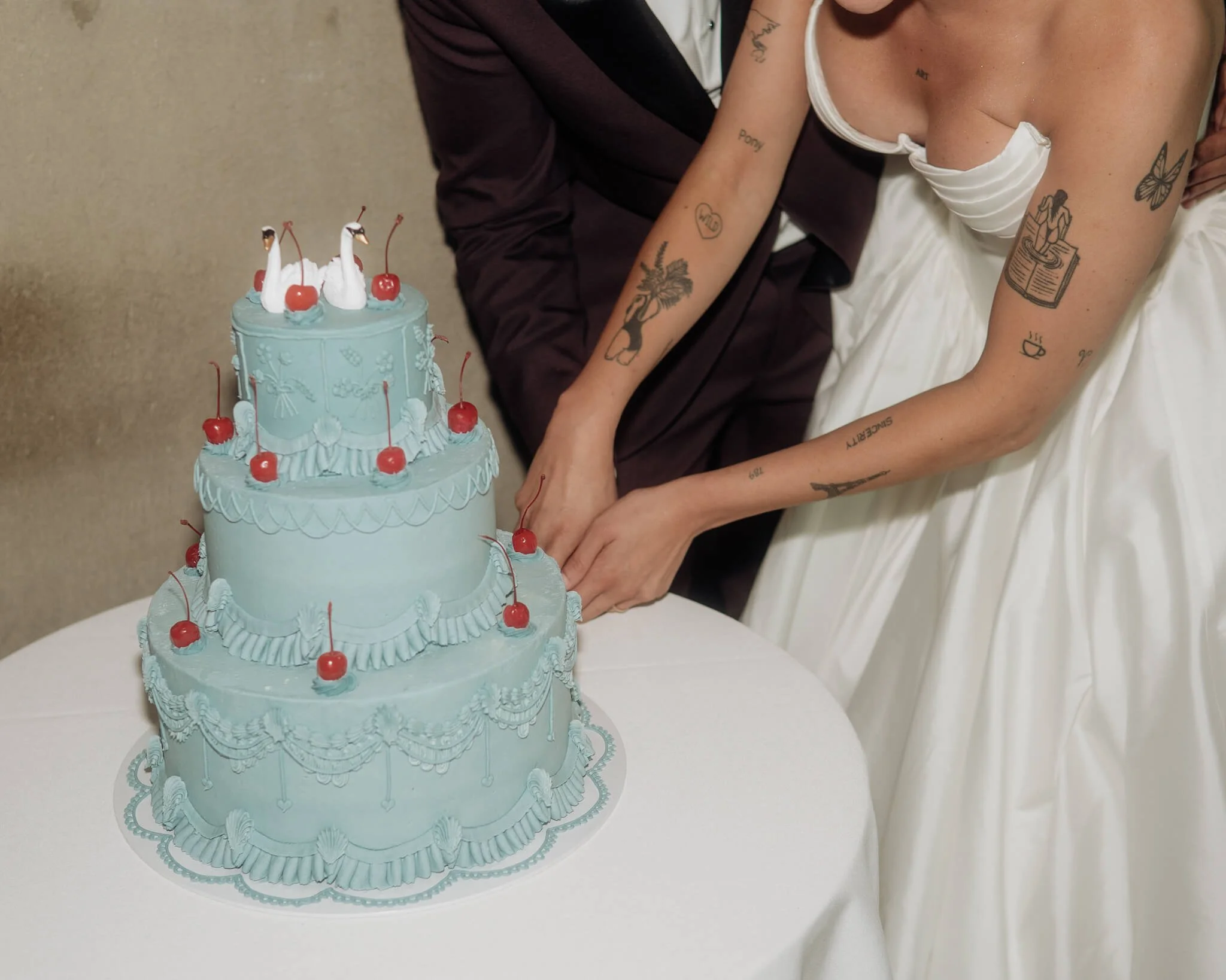 A couple dressed in wedding attire cutting a light blue, three-tiered wedding cake decorated with red cherries and swan cake toppers. The bride has tattoos on her arms, and the groom is wearing a dark suit.