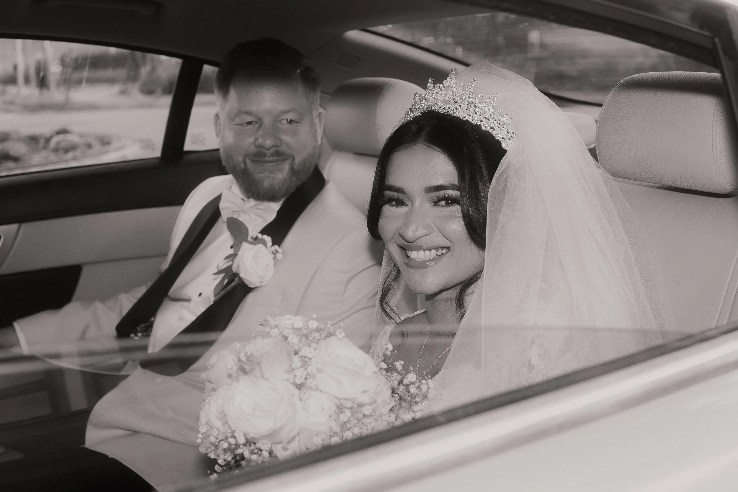 A smiling bride and groom sitting in a car on their wedding day, with the bride wearing a tiara and veil, holding a bouquet, and the groom in a tuxedo with a boutonniere.