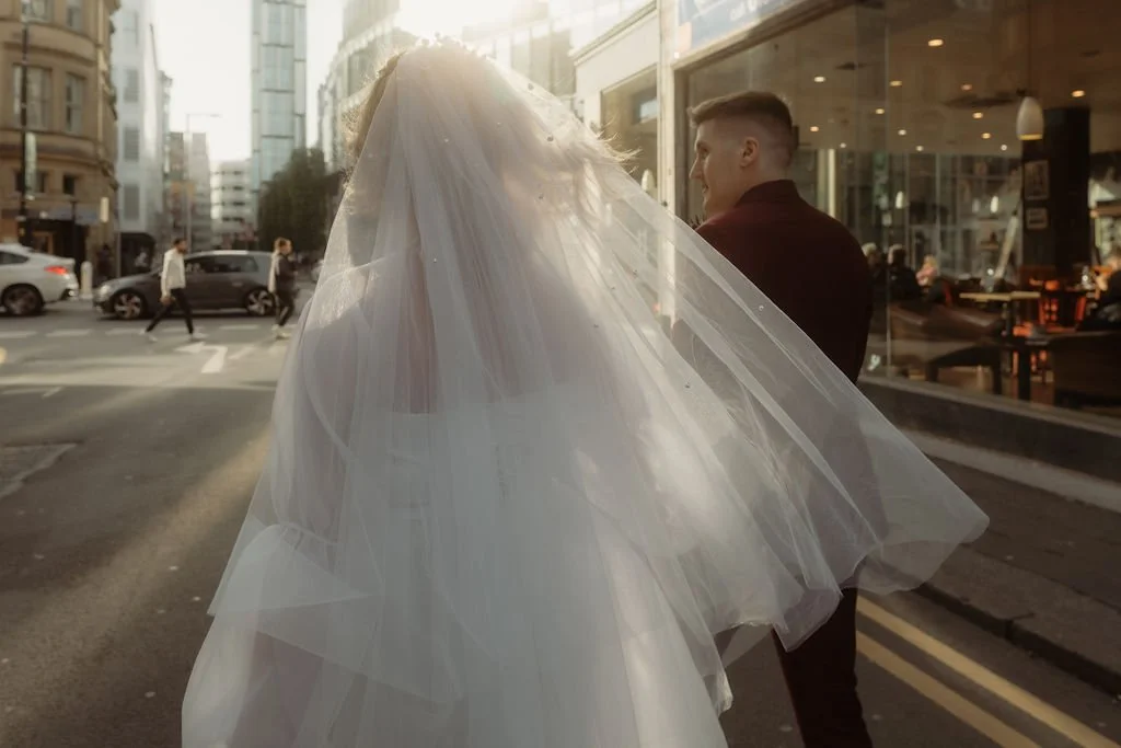 A bride in a white wedding dress with a veil walking on a city street at sunset, accompanied by a man in a dark jacket.