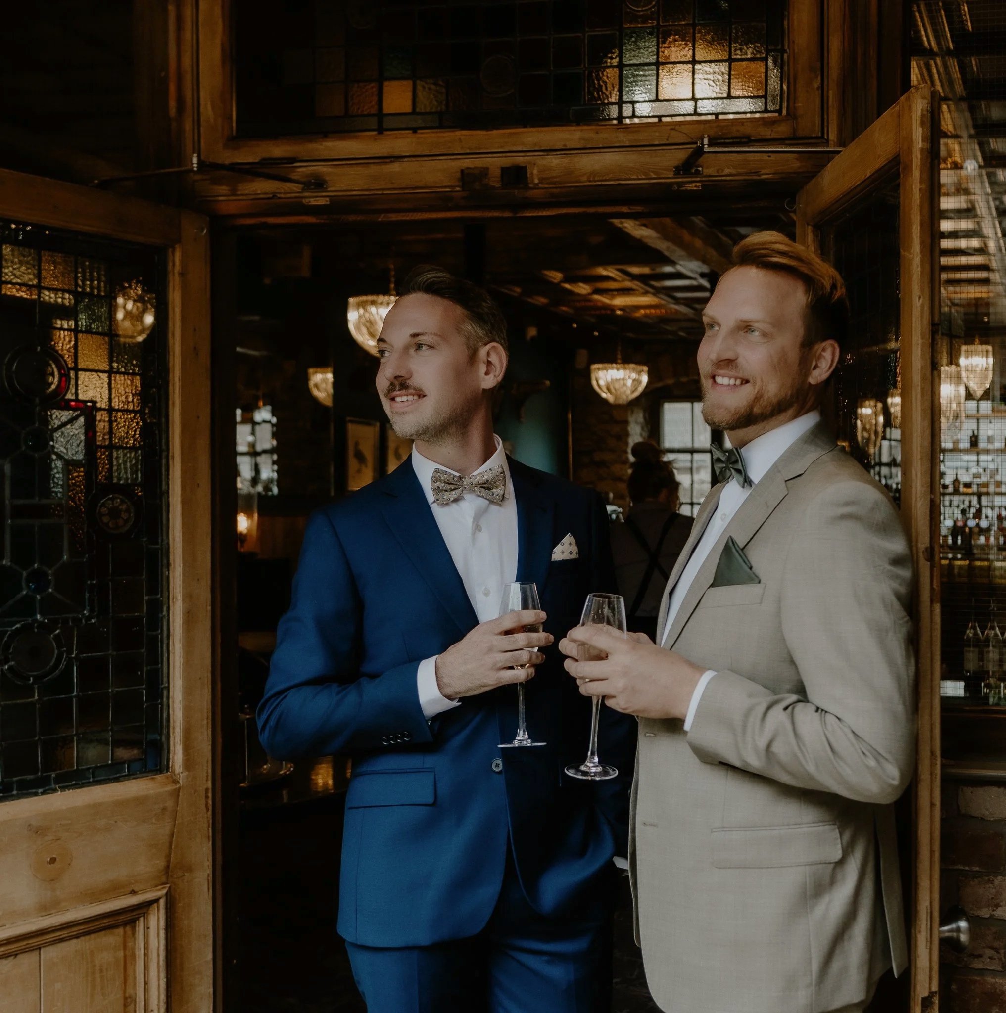 Two men in suits holding glasses of champagne at a formal event in a rustic, warmly lit venue.