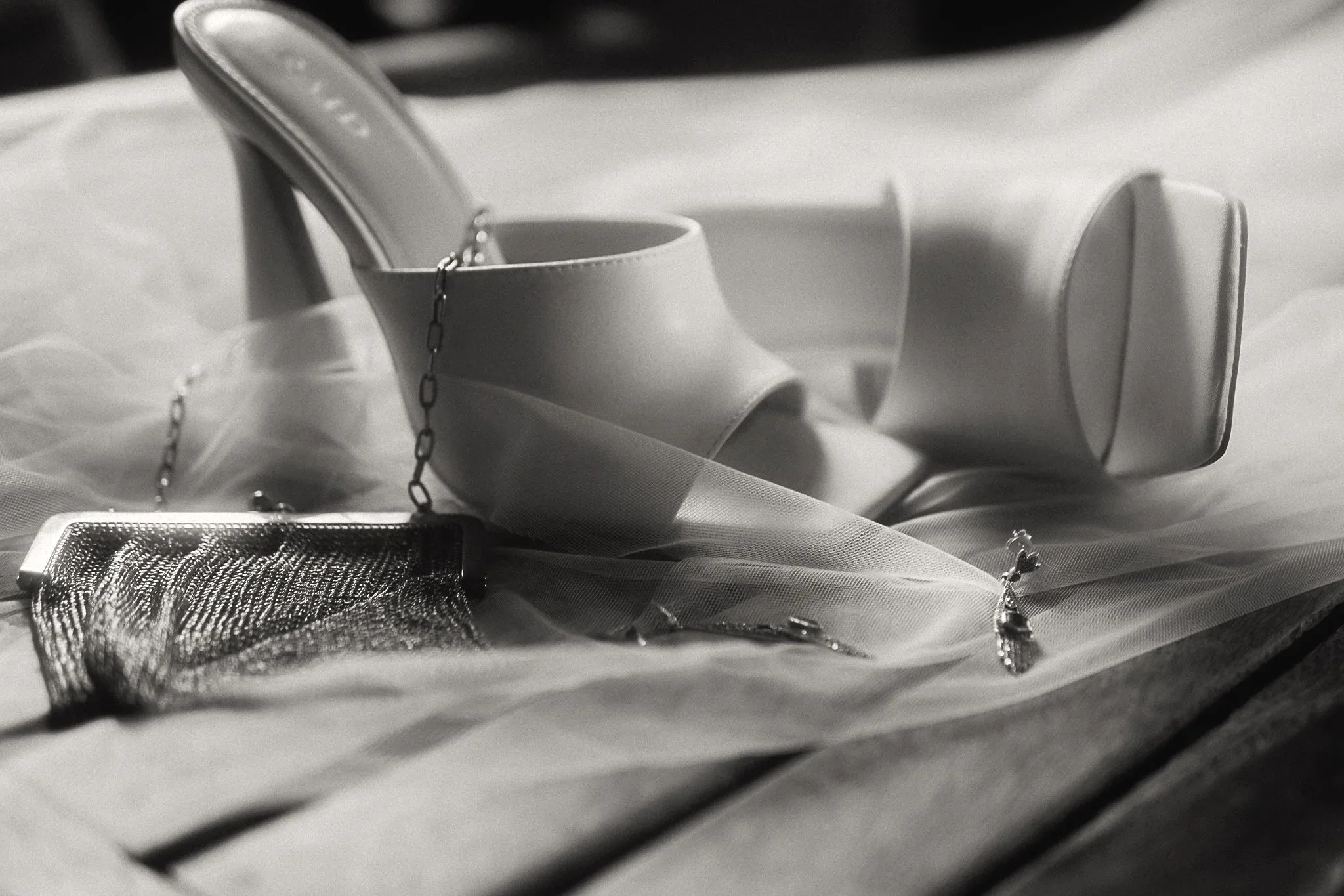 A pair of elegant high-heeled shoes, a small mesh purse, and jewelry lie on a soft, sheer fabric surface, captured in black-and-white.