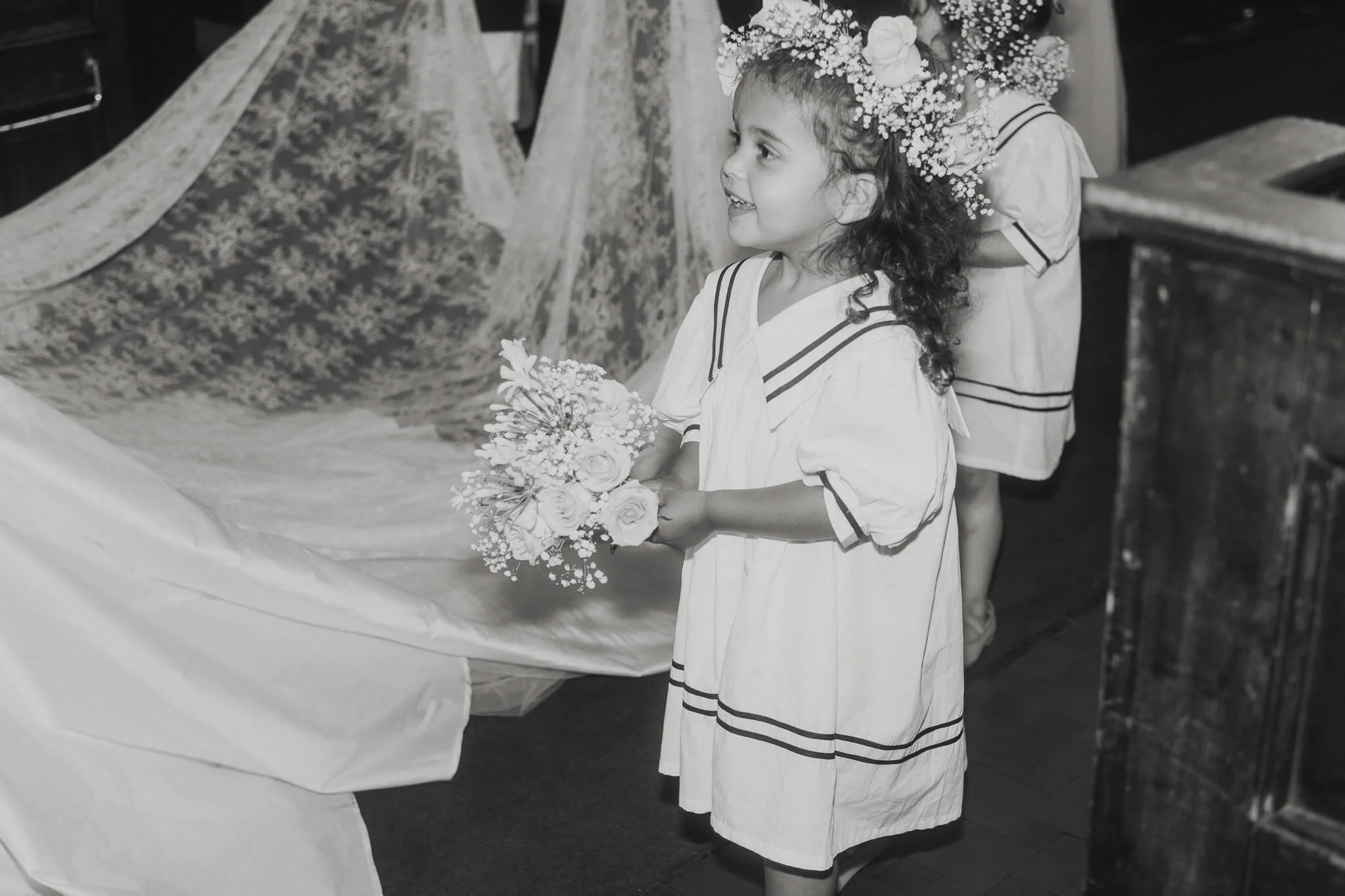 A young girl with curly hair wearing a dress with stripe details, holding a bouquet of flowers, and wearing a flower crown, smiling in a black and white photo at a wedding ceremony.