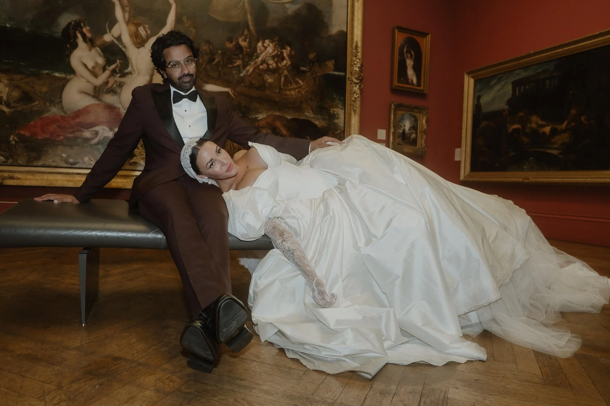 A man in a tuxedo and a woman in a white wedding dress posing in an art gallery with framed paintings in the background.