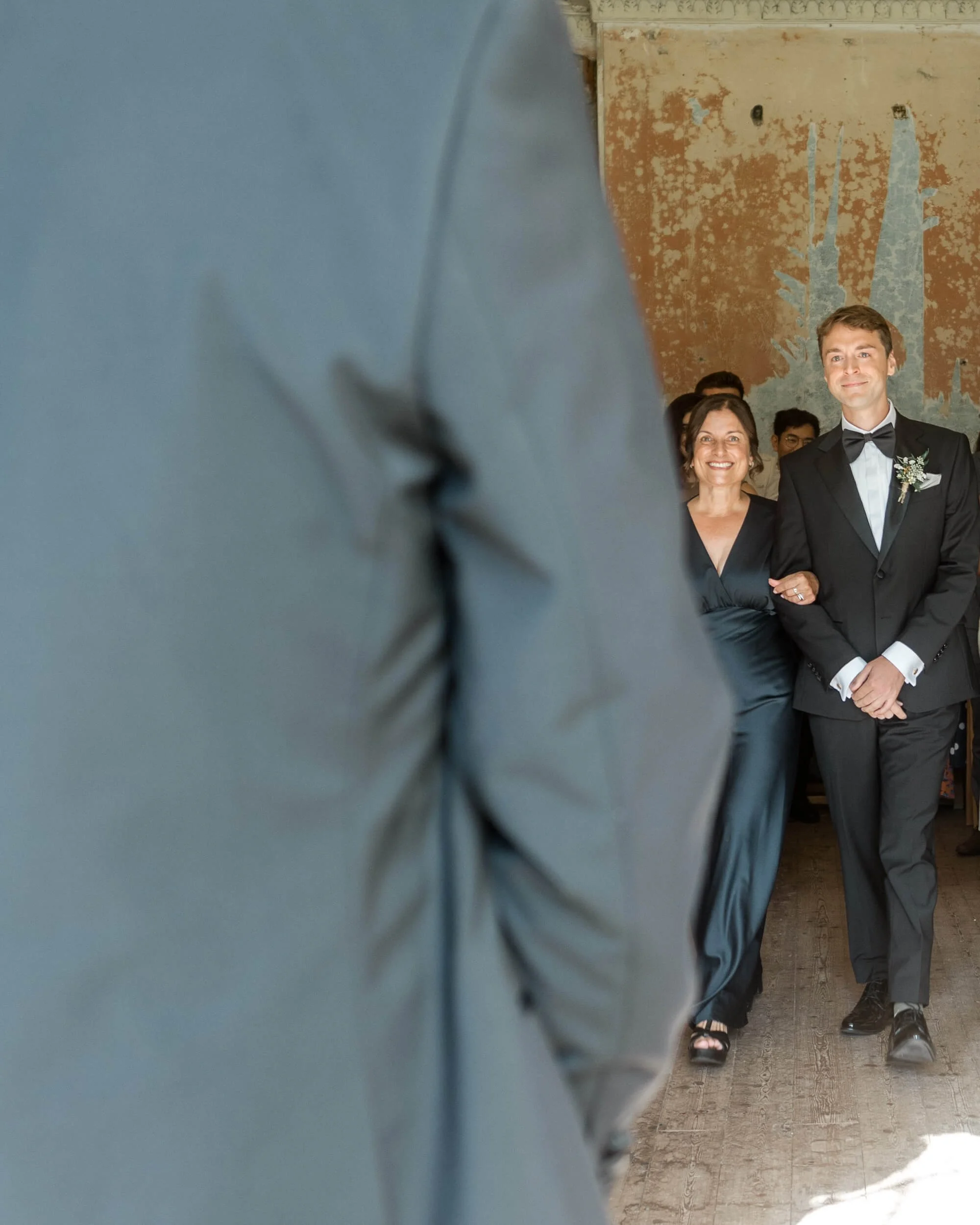 A young man in a tuxedo walking down an aisle with a mum, with several people standing behind them, at a wedding ceremony.