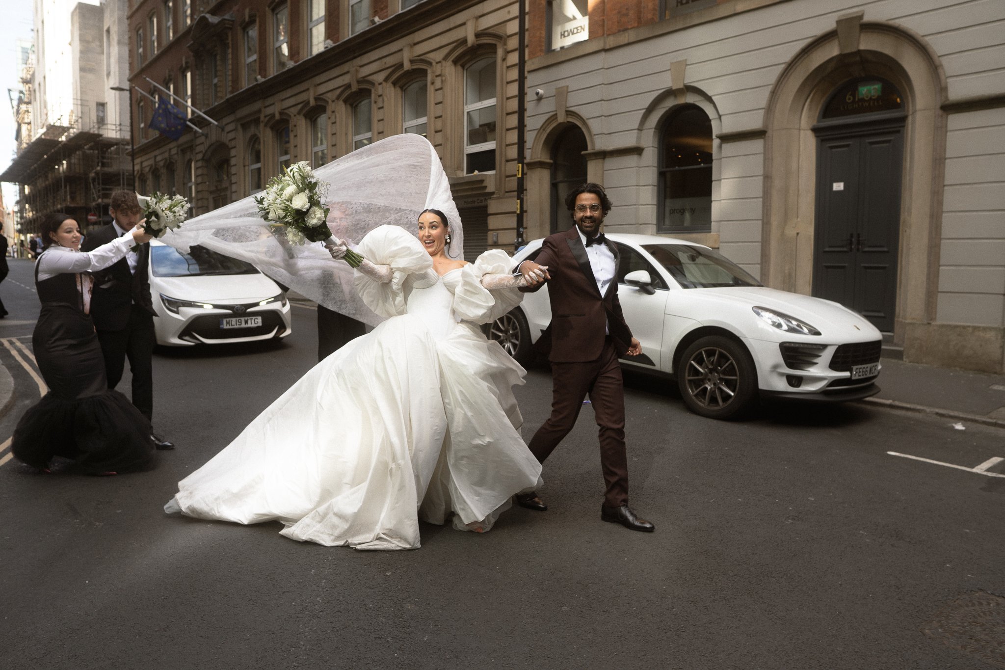A bride and groom joyfully walk hand-in-hand on a city street, surrounded by their wedding party.