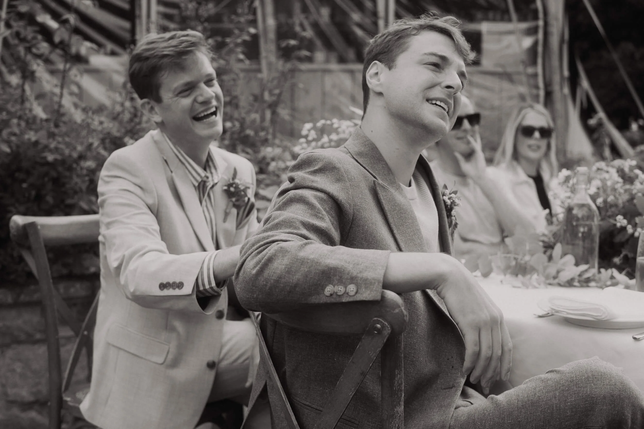 People sitting at a table outdoors, smiling and laughing, in a candid moment, with blurred greenery in the background.