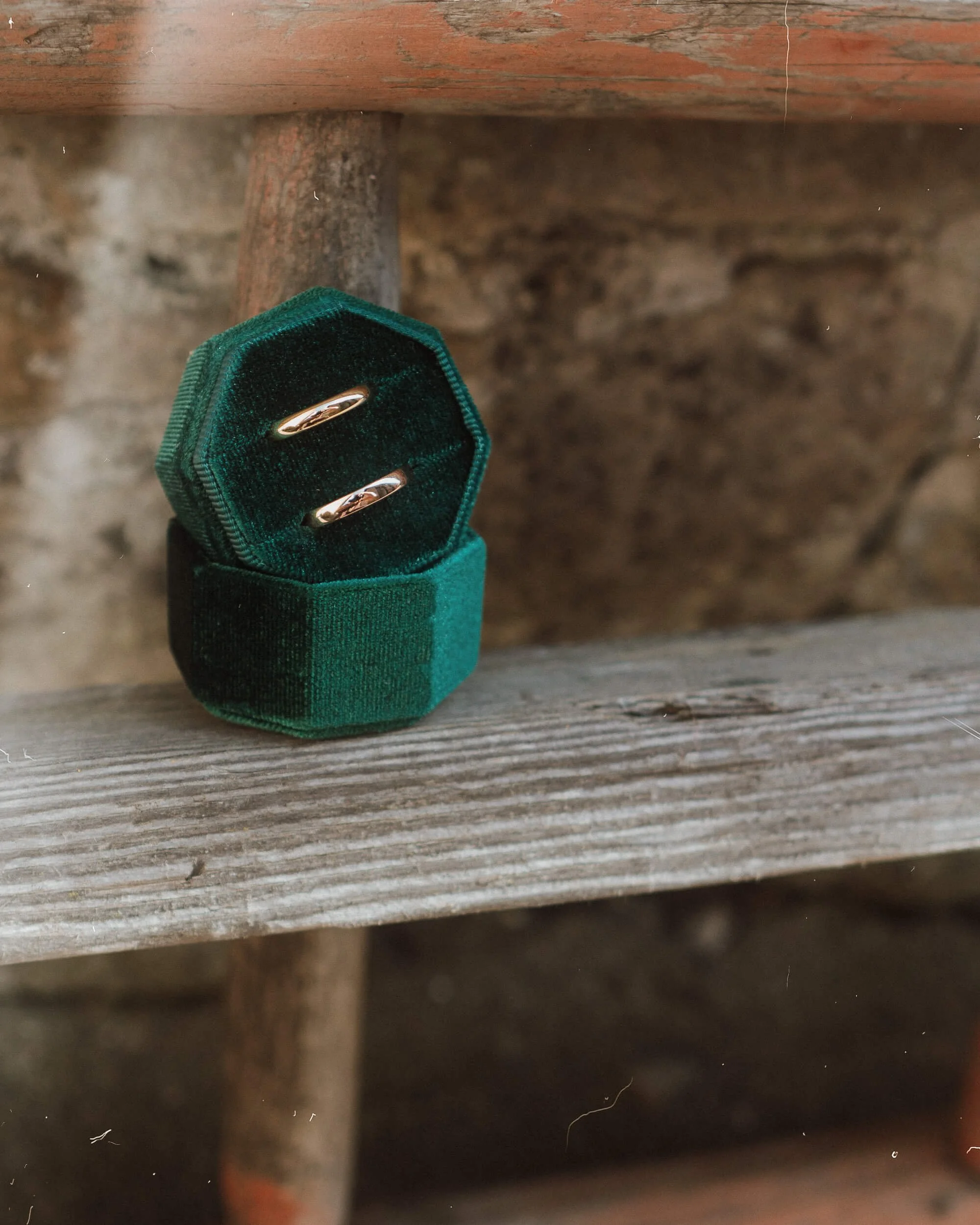 A pair of gold wedding rings in a green velvet ring box on a wooden surface with a stone wall background.