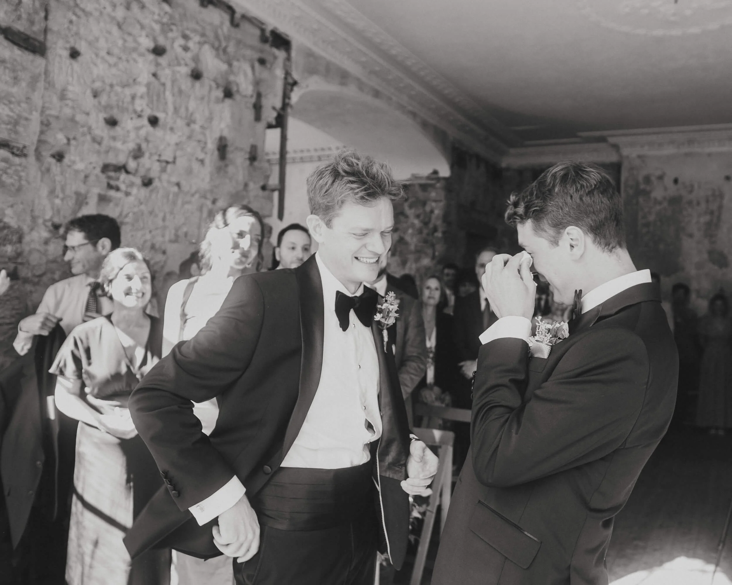 Two men in tuxedos are standing face-to-face, smiling and holding hands during a wedding ceremony. Several guests are in the background in a rustic indoor setting.
