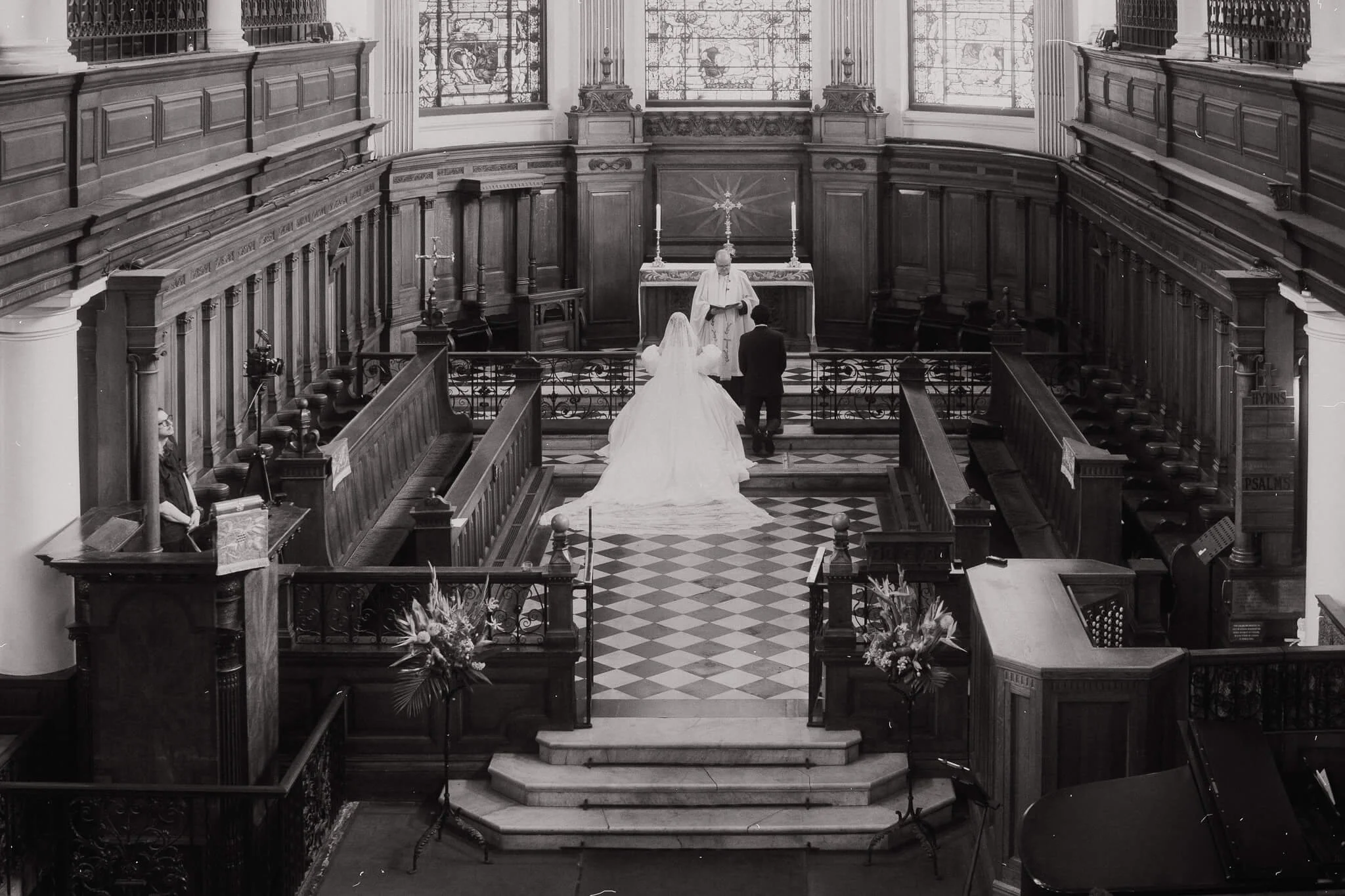 A black and white photo of a wedding ceremony inside a church with wooden pews, stained glass windows, and religious statues. The bride and groom are kneeling before a priest at the altar.
