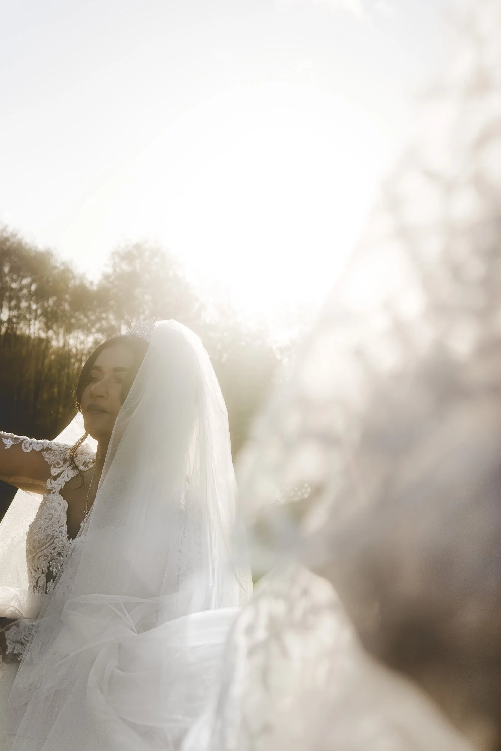 A bride in a lace wedding dress and veil sits outdoors in sunlight, with trees in the background and a large floral arrangement in the foreground.