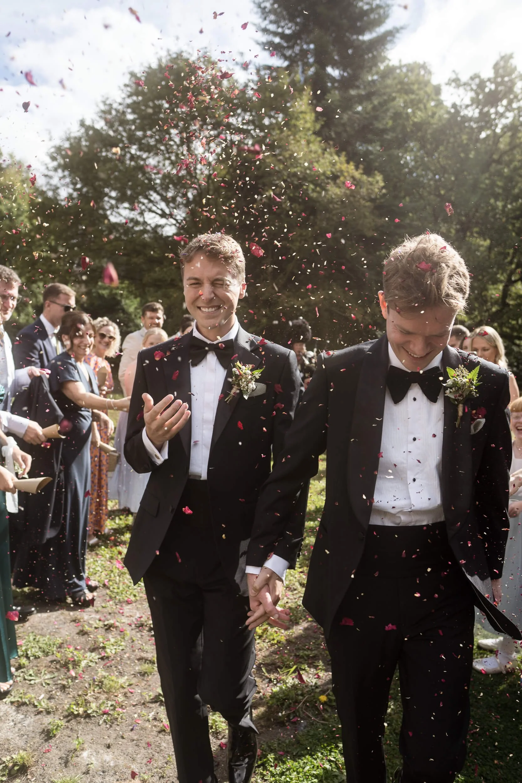 Two men in tuxedos celebrating at a wedding, holding hands and smiling as flower petals are thrown in the air.