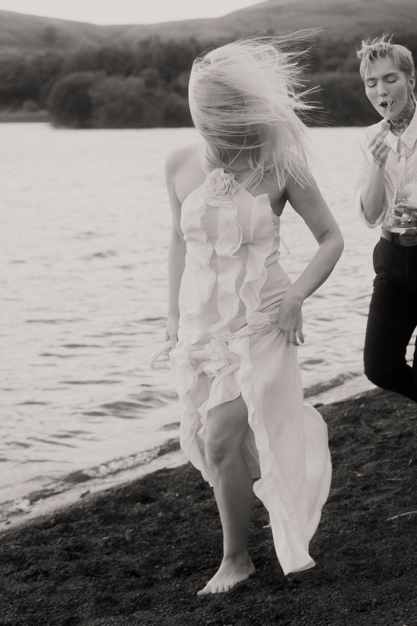 A woman in a ruffled dress is walking barefoot on a beach near water, with wind blowing her blonde hair. Next to her, a person with short hair and a tattoo on their neck is eating. The scene appears to be outdoors near a lake or sea with hills in the