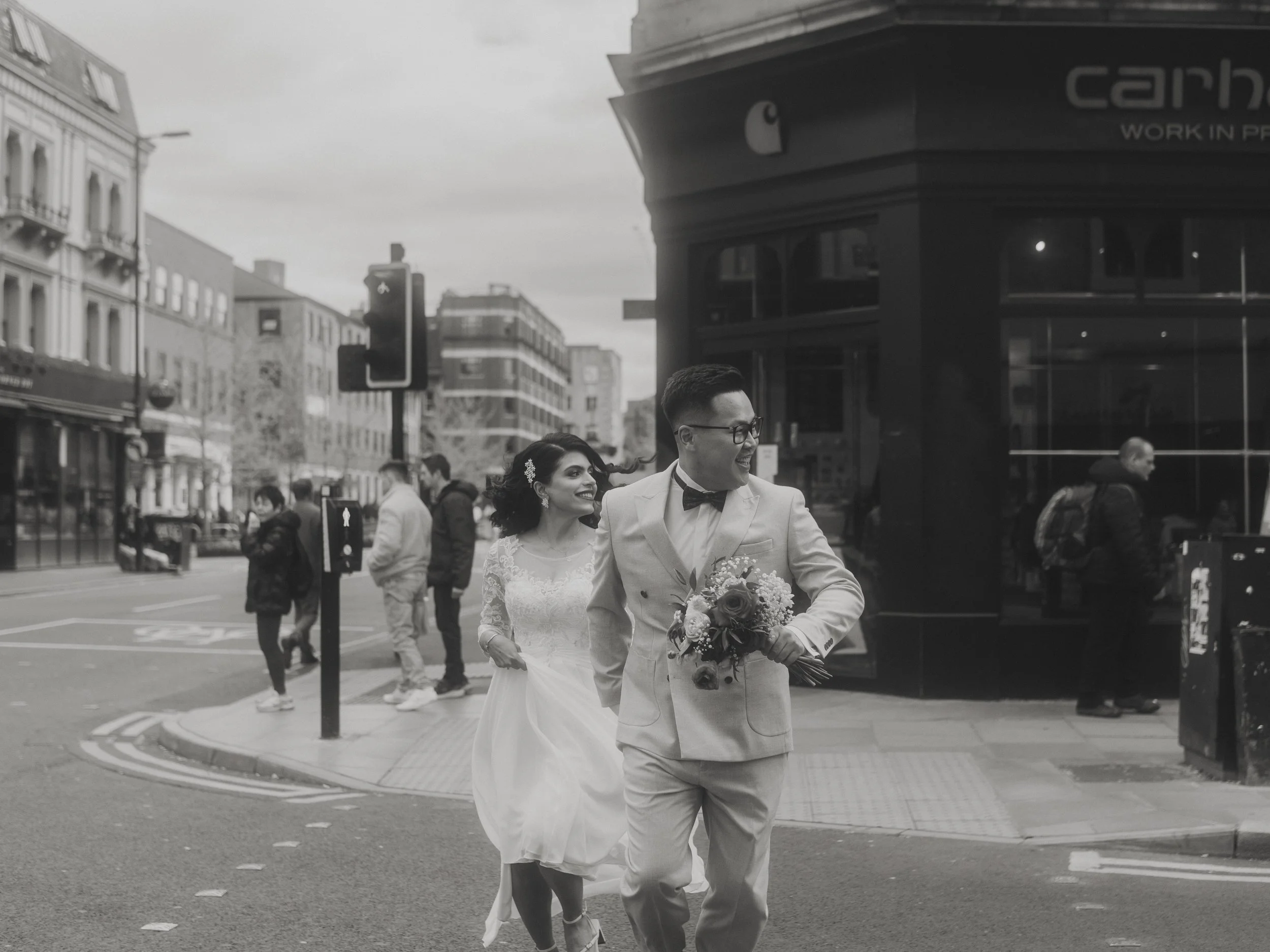 A black and white photo of a newlywed couple dressed in wedding attire, walking on a city street corner with pedestrians and buildings in the background.