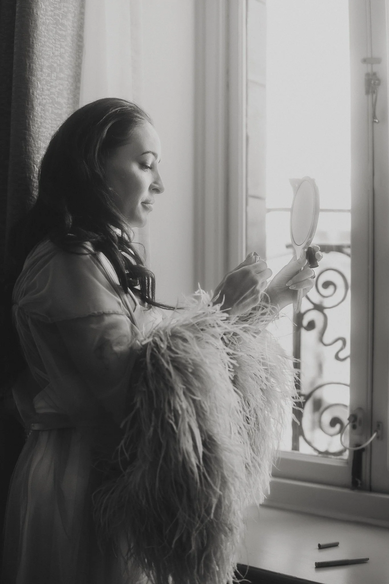 Woman applying makeup while looking in a mirror by a window.