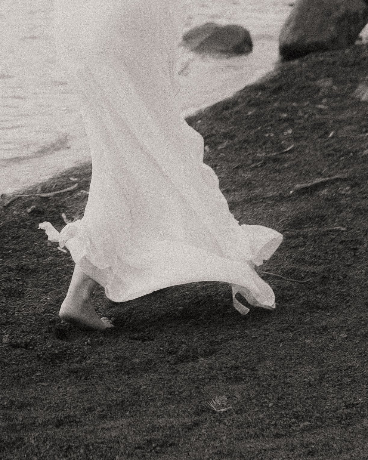 A person is walking barefoot on dark sand near the water, wearing a flowing white dress.