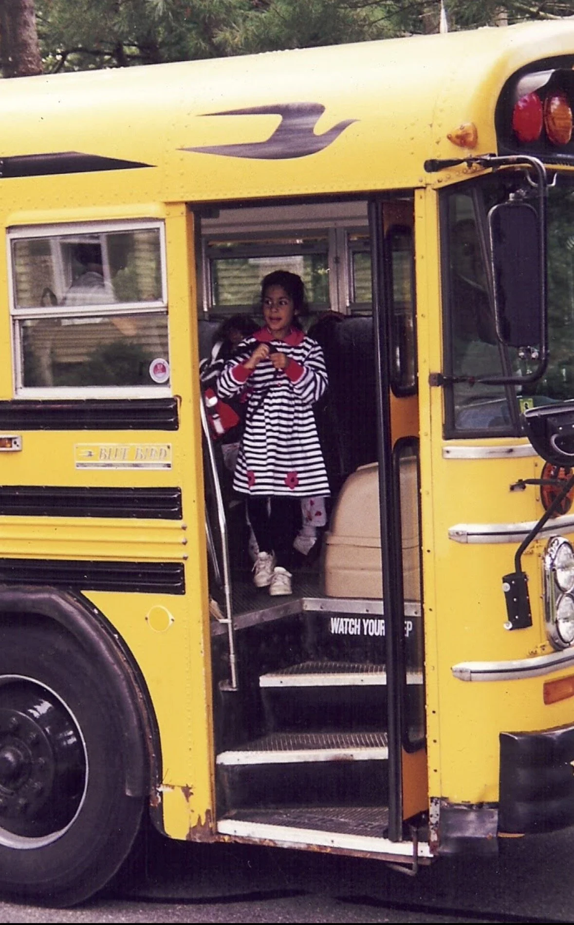 a 7-year-old Shivani stands at the top of the steps, about to descend from a yellow school bus. she's wearing a black and white striped dress with red flowers and holding a black, red, and white backpack.