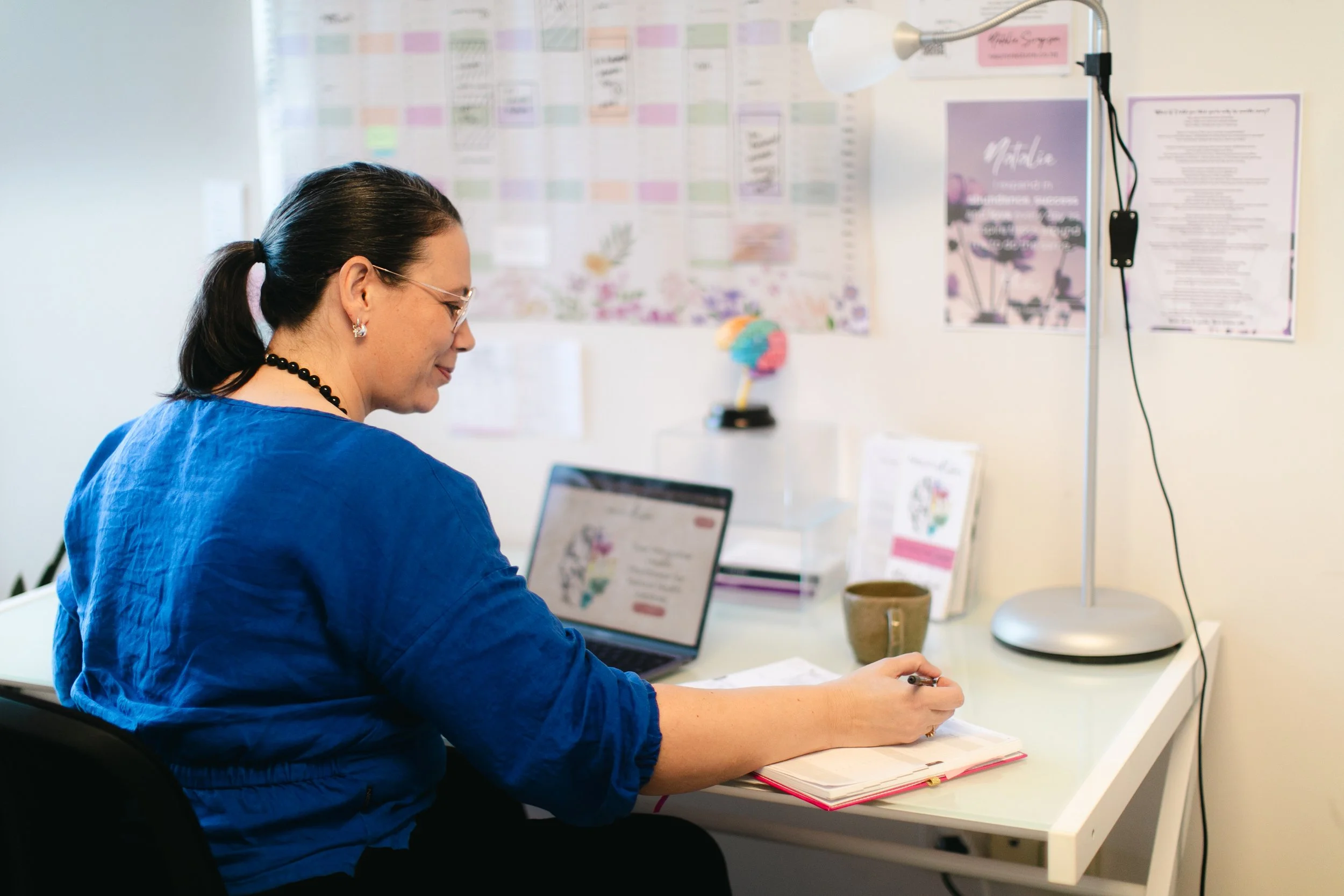 A woman with dark hair tied back, wearing glasses, a blue shirt, and earrings, sitting at a white desk with a laptop, notebook, and a cup, working in an office with a wall covered in colorful charts and posters, a tall white lamp, and a window.