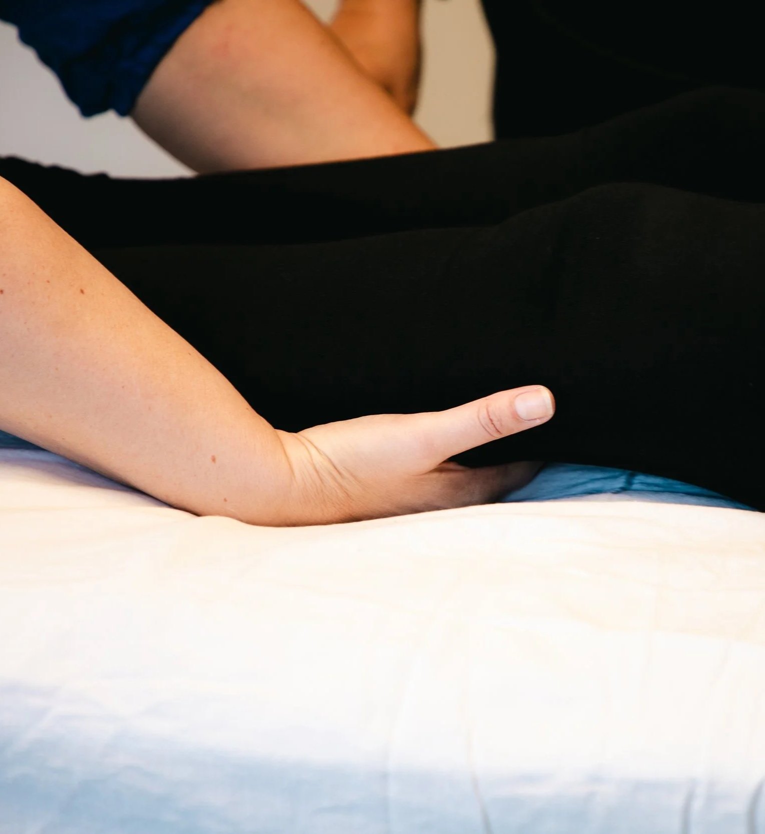 A person lying on a bed with their arm resting on the mattress and their finger extended, wearing a black shirt and blue jeans.