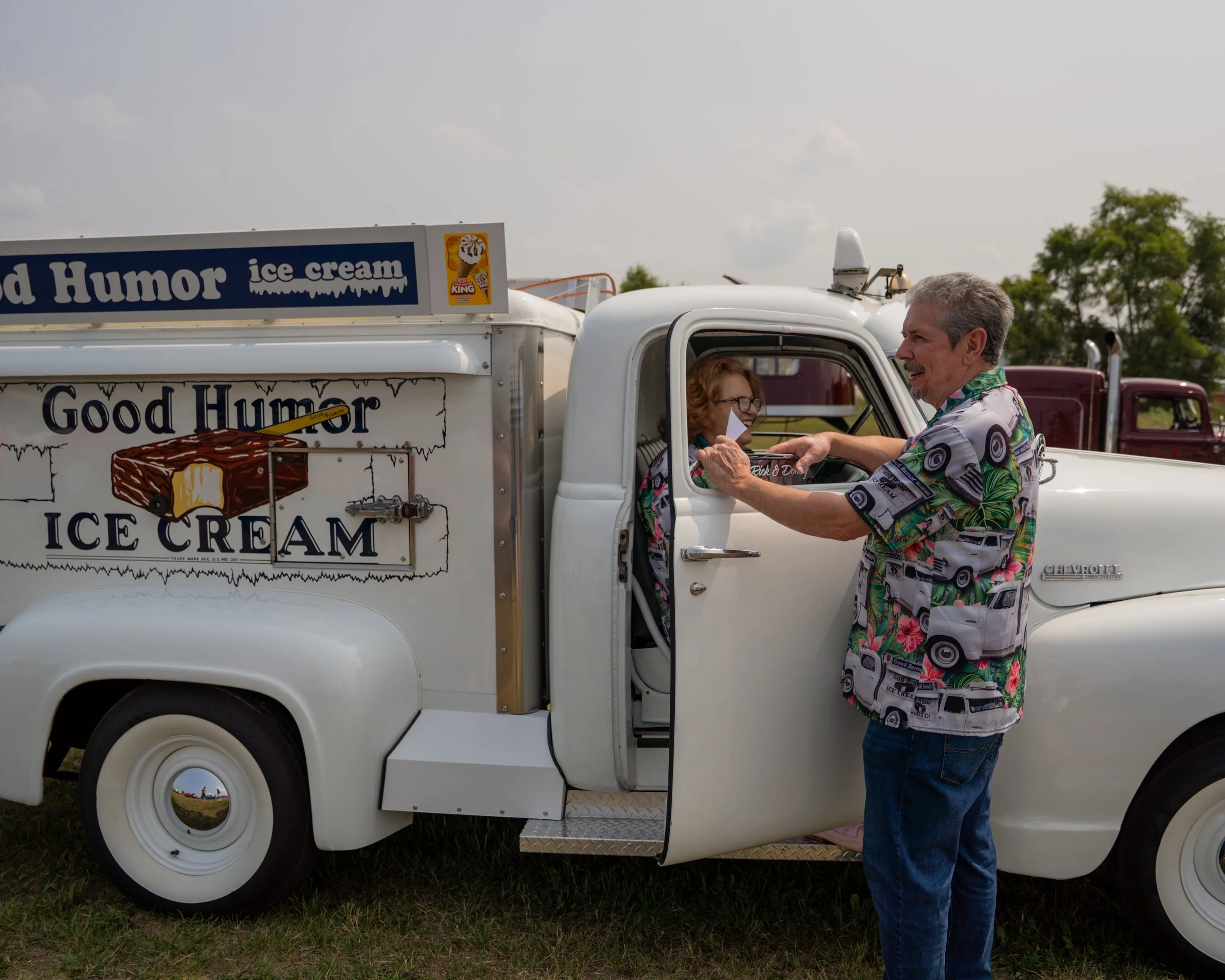 A man in vibrant, colorful clothing gives money to a woman sitting inside a vintage ice cream truck at an outdoor event.