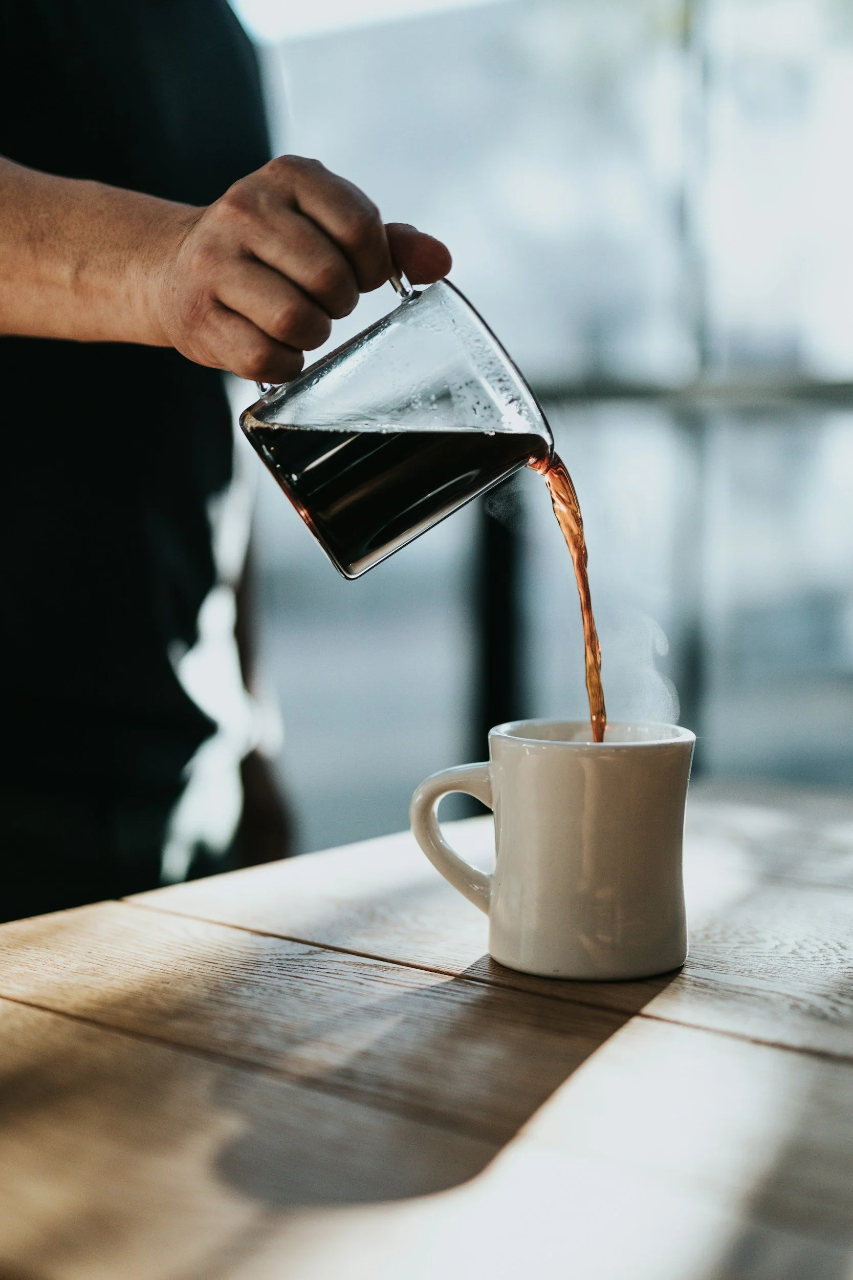 Person pouring hot black coffee from a glass carafe into a white ceramic mug on a wooden table.