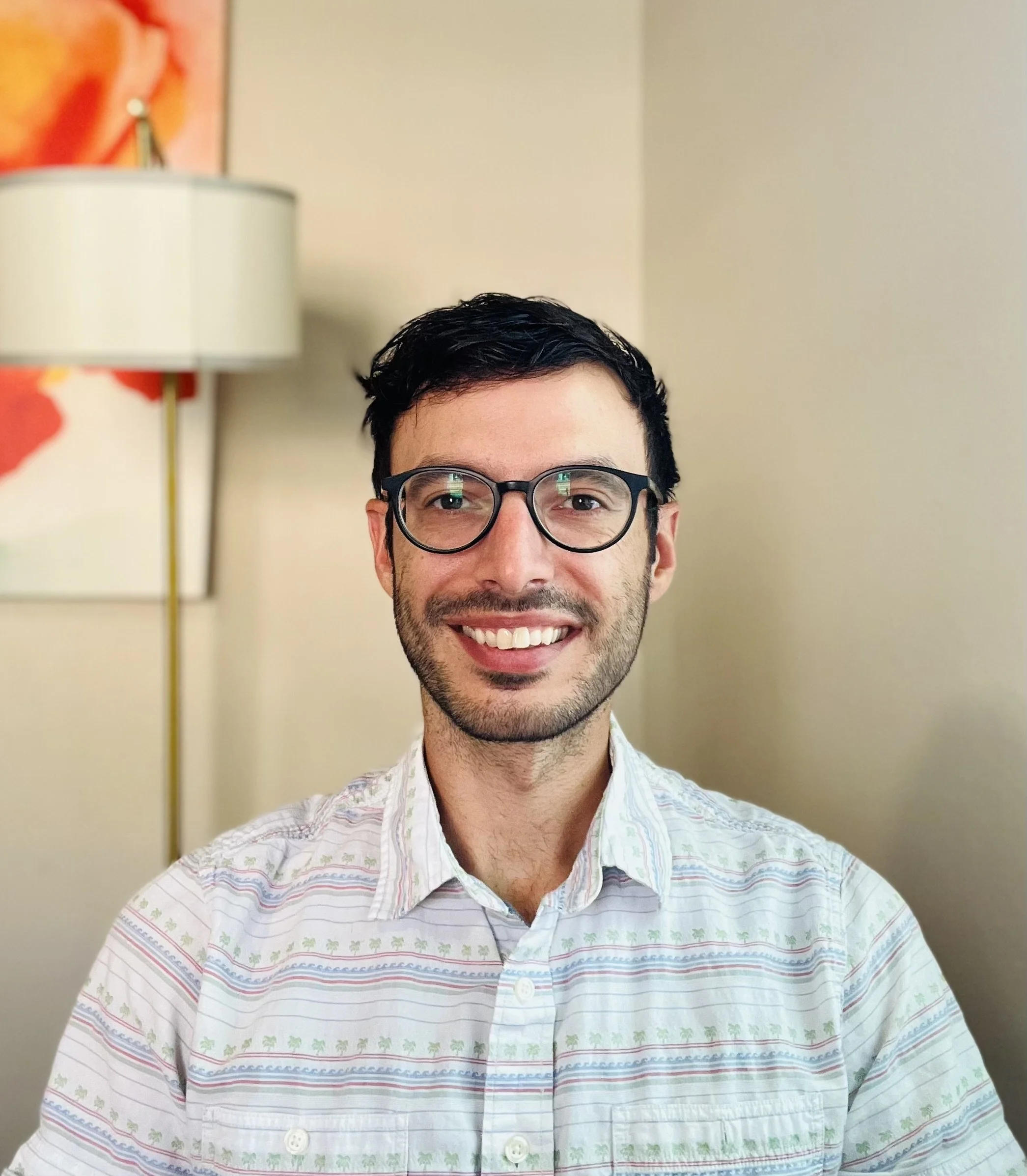 A young man with glasses smiling indoors, with a lamp and abstract artwork in the background.