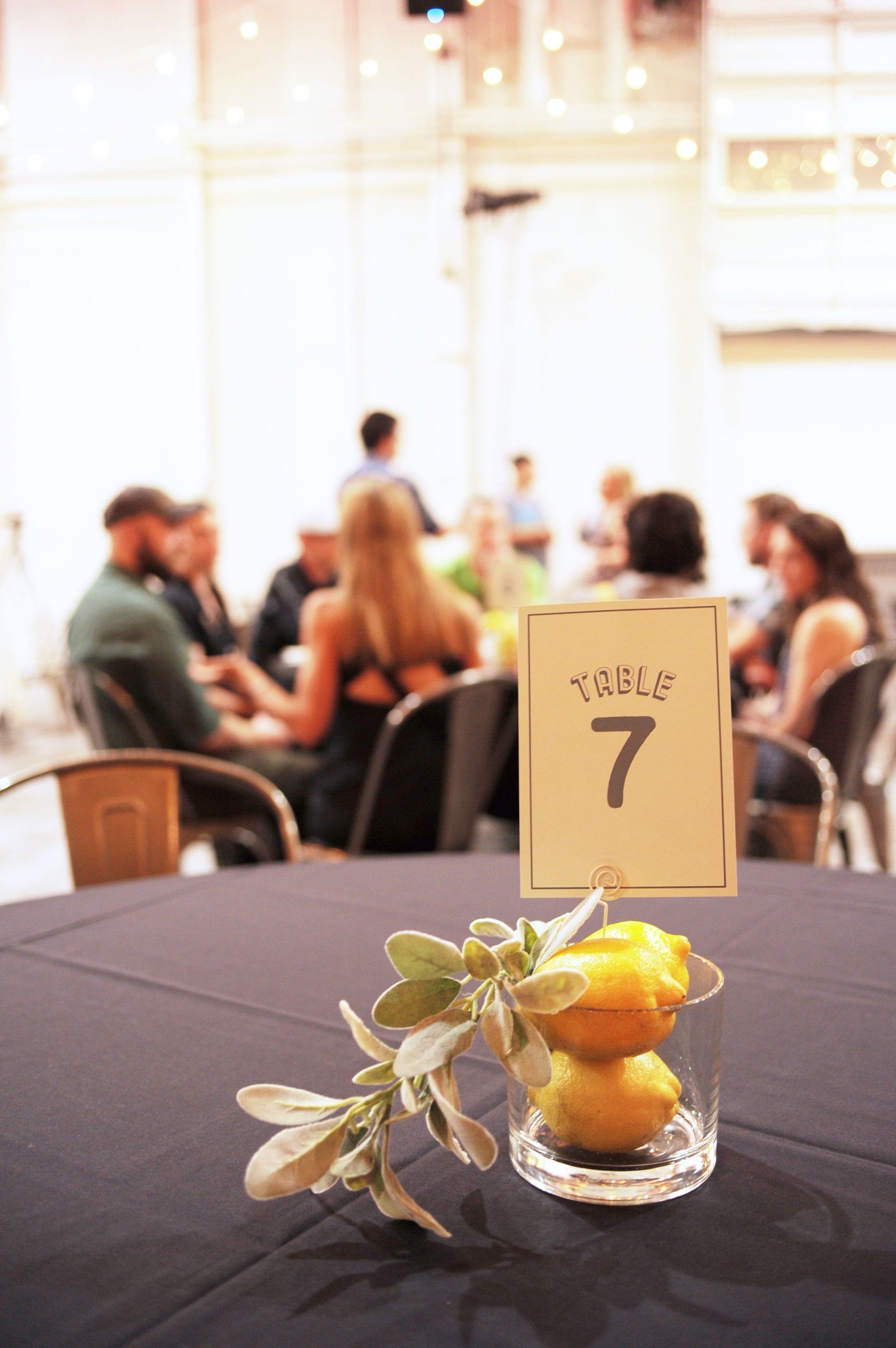 A table with a sign labeled "Table 7" and a glass containing lemons and a decorative sprig of greenery, set in a venue with blurred people in the background.