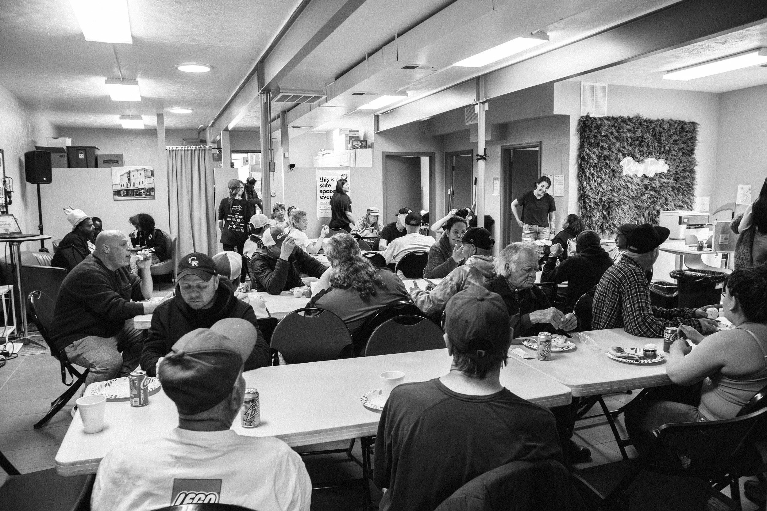 Black and white photo of a man wearing glasses, a face mask, a beanie, and a jacket, standing and facing two women who are also wearing masks and jackets, in an indoor setting.