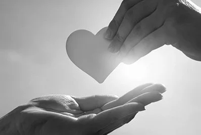 Two hands exchanging a paper heart in black and white, symbolizing love or care.