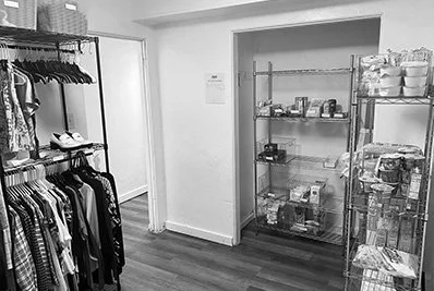 A small pantry or storage room with metal shelving holding packaged food items and snacks, alongside a clothing rack with hanging shirts and dresses, in a room with wooden flooring and white walls.