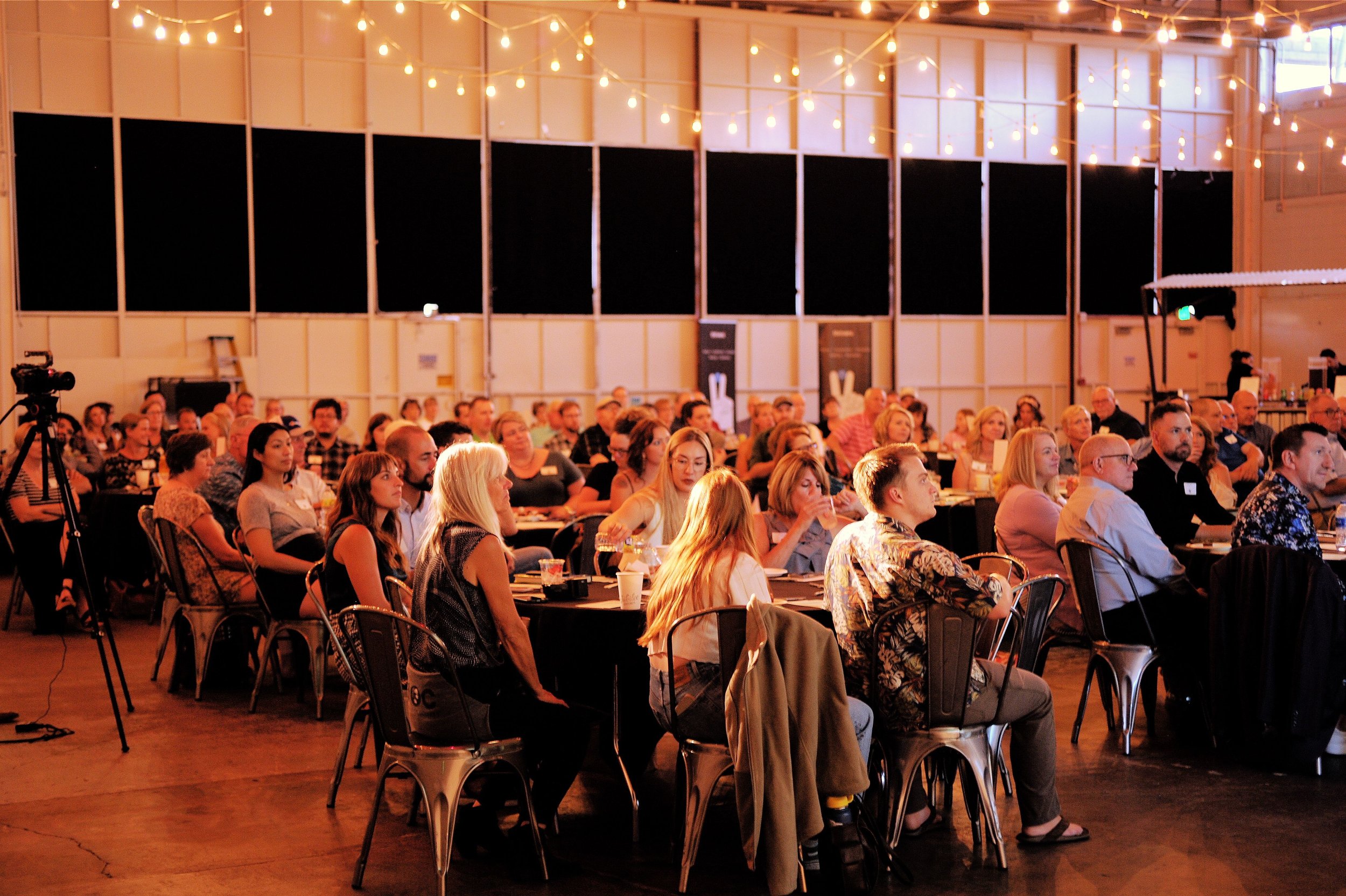 Audience attending a conference or seminar in a large hall with string lights hanging from the ceiling, seated at round tables.