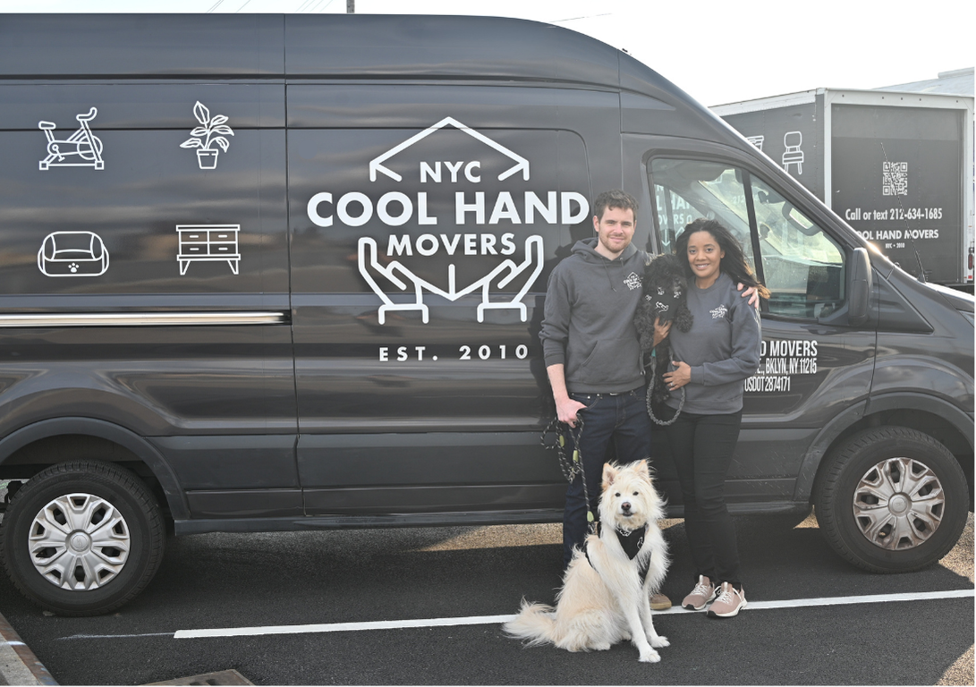Two people standing in front of a black NYC Cool Hand Movers van, both wearing gray hoodies with the company's logo, holding two dogs.