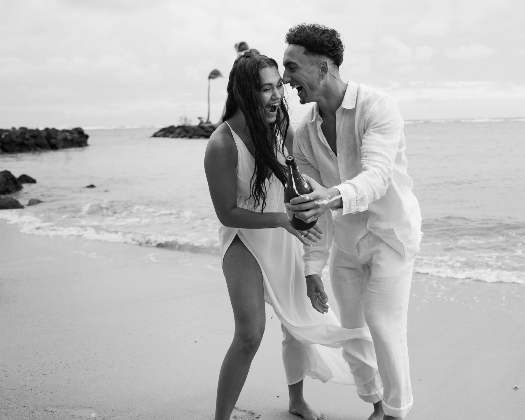 A black and white photo of a couple laughing and enjoying themselves at the beach, holding a bottle of beer.