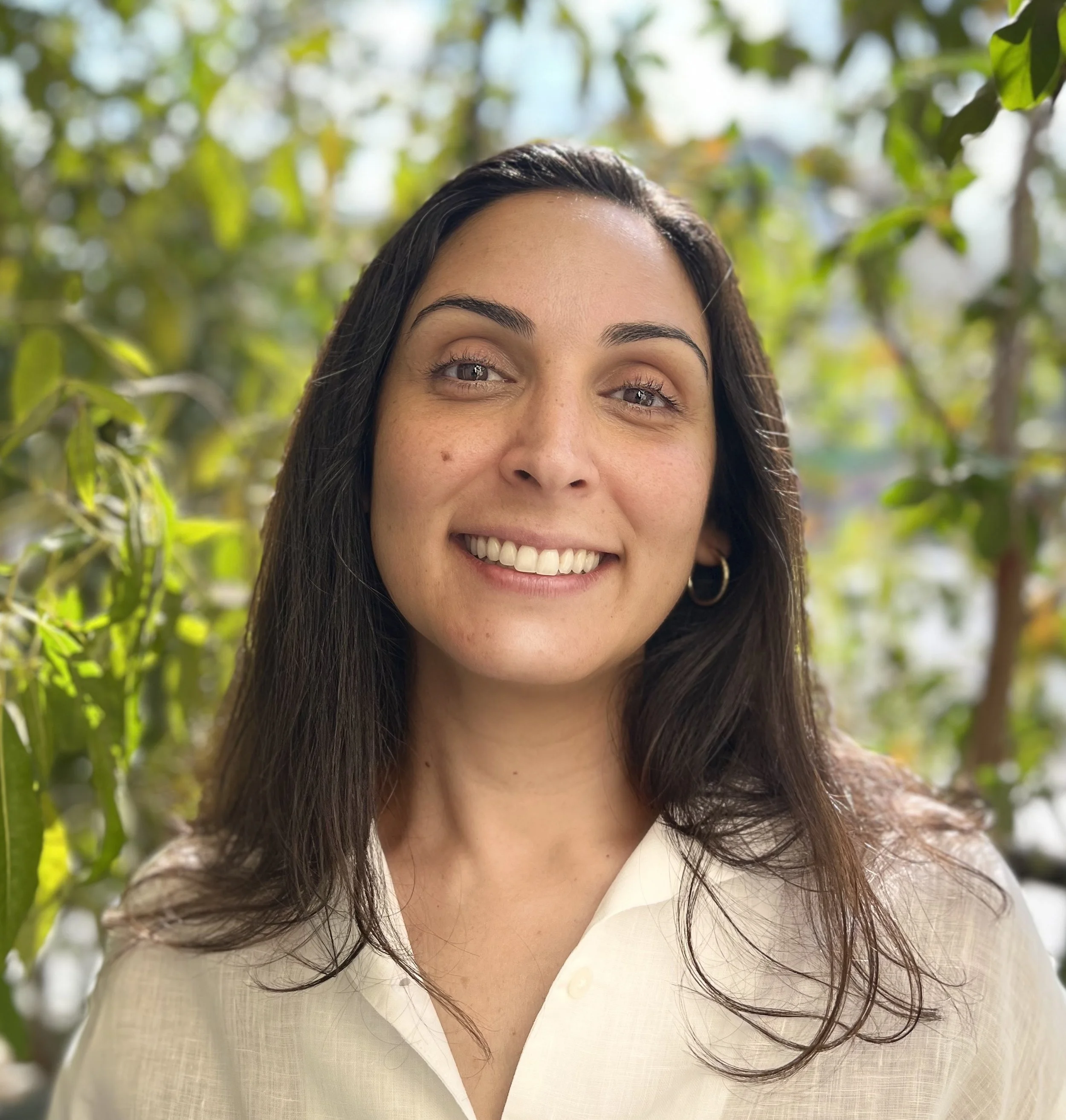 A smiling woman with dark hair and hoop earrings stands in front of green leafy plants.