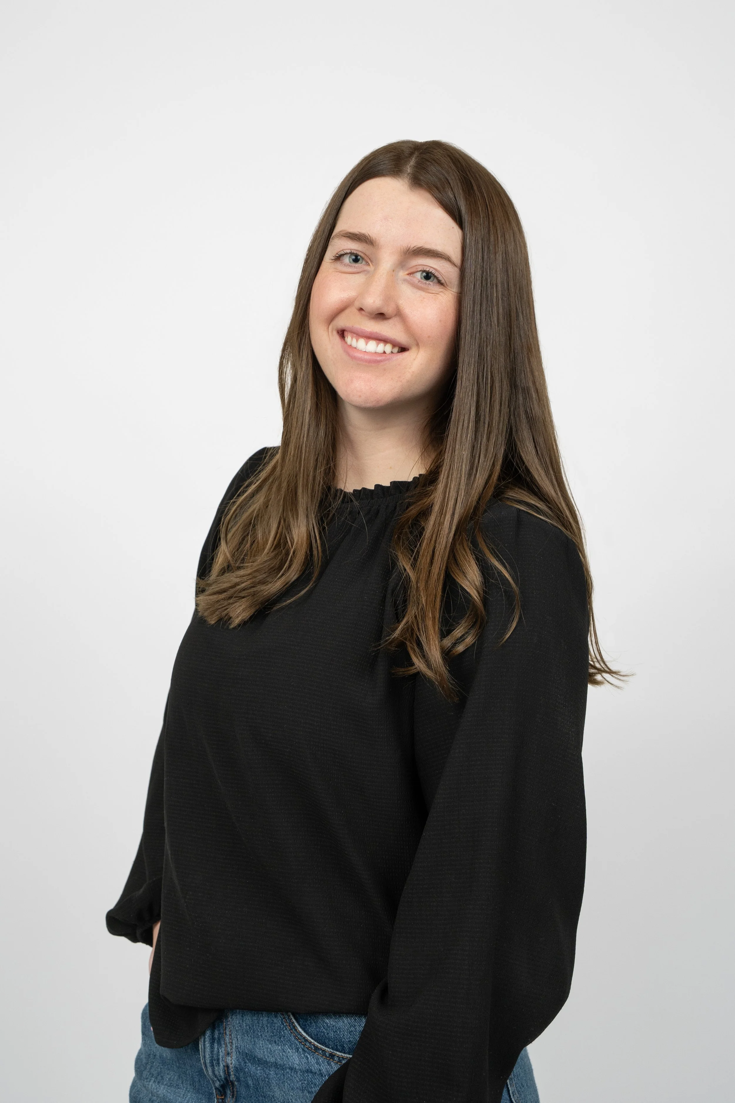 A young woman with long brown hair, smiling, wearing a black blouse and blue jeans, standing against a plain white background.