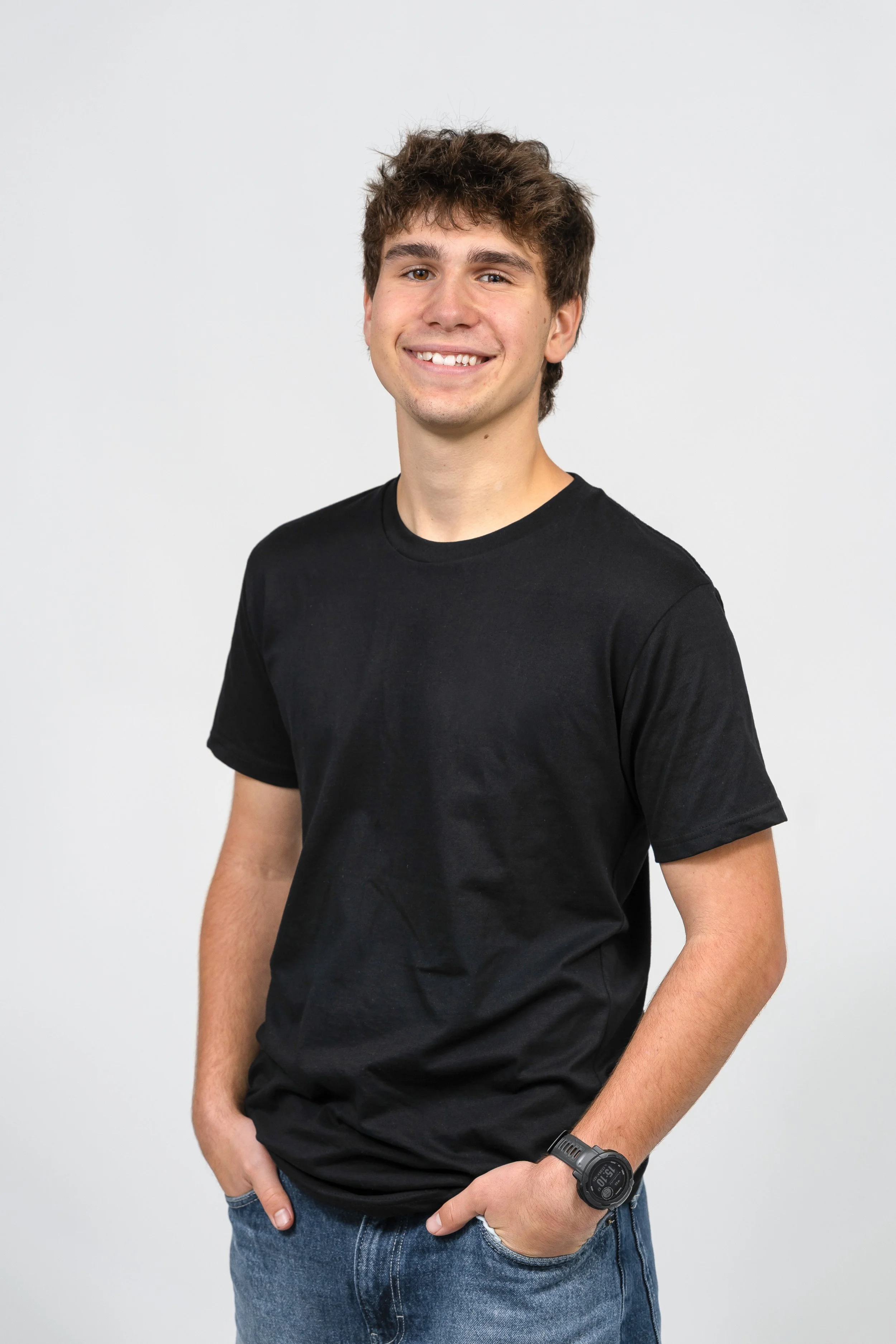 A young man with brown curly hair wearing a black t-shirt and blue jeans, smiling with hands in pockets, standing against a plain white background.