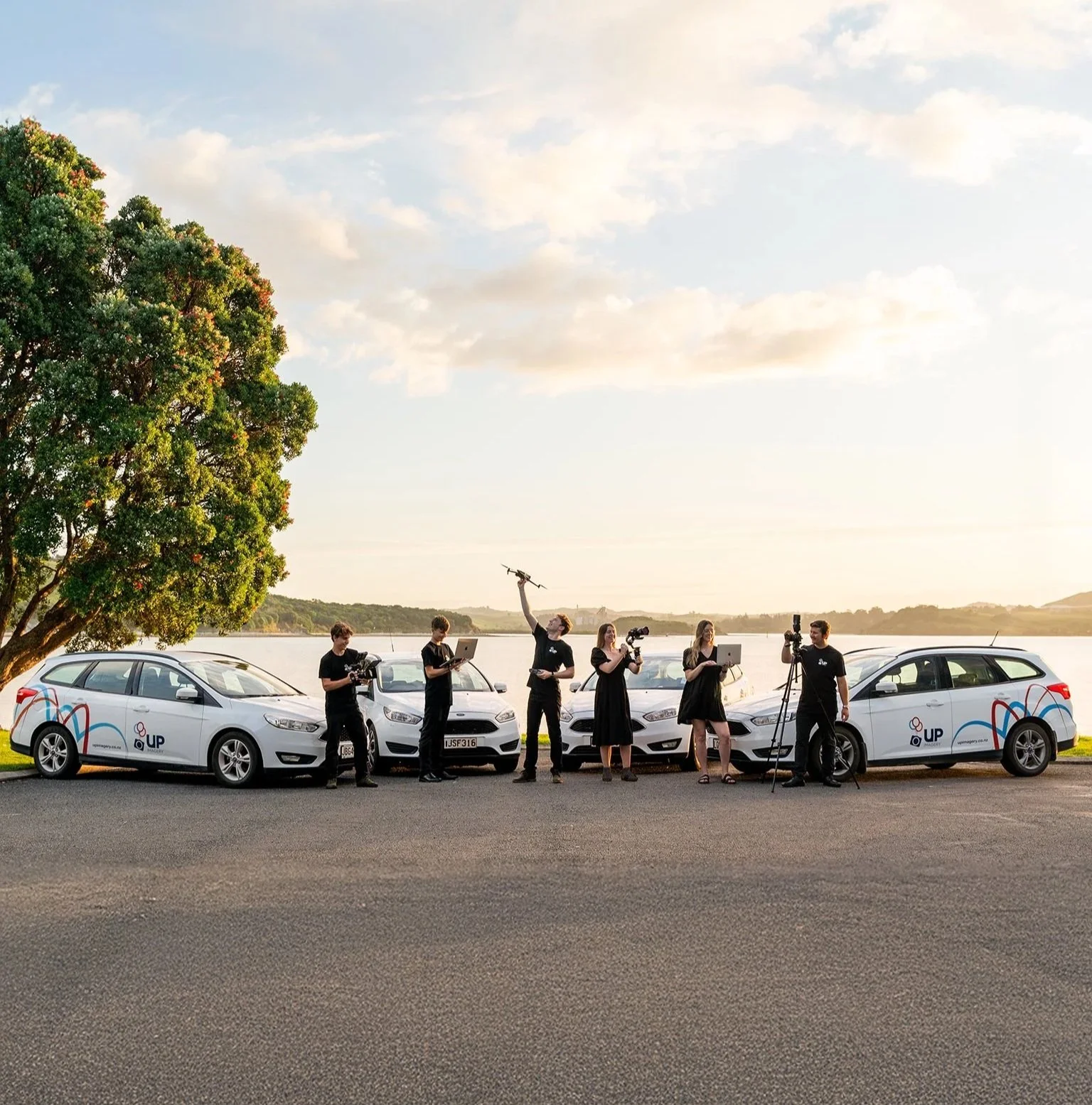 Group of six people, some with cameras, standing in front of white cars by a lake, with a tree on the left, during sunset.