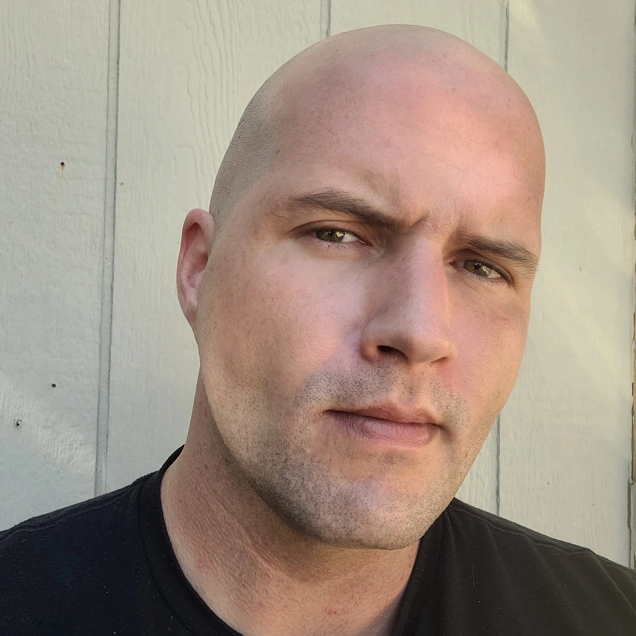 image of the artist dopeula, a bald man with light skin and green eyes, wearing a black shirt, standing outside in front of a light-colored wooden wall.