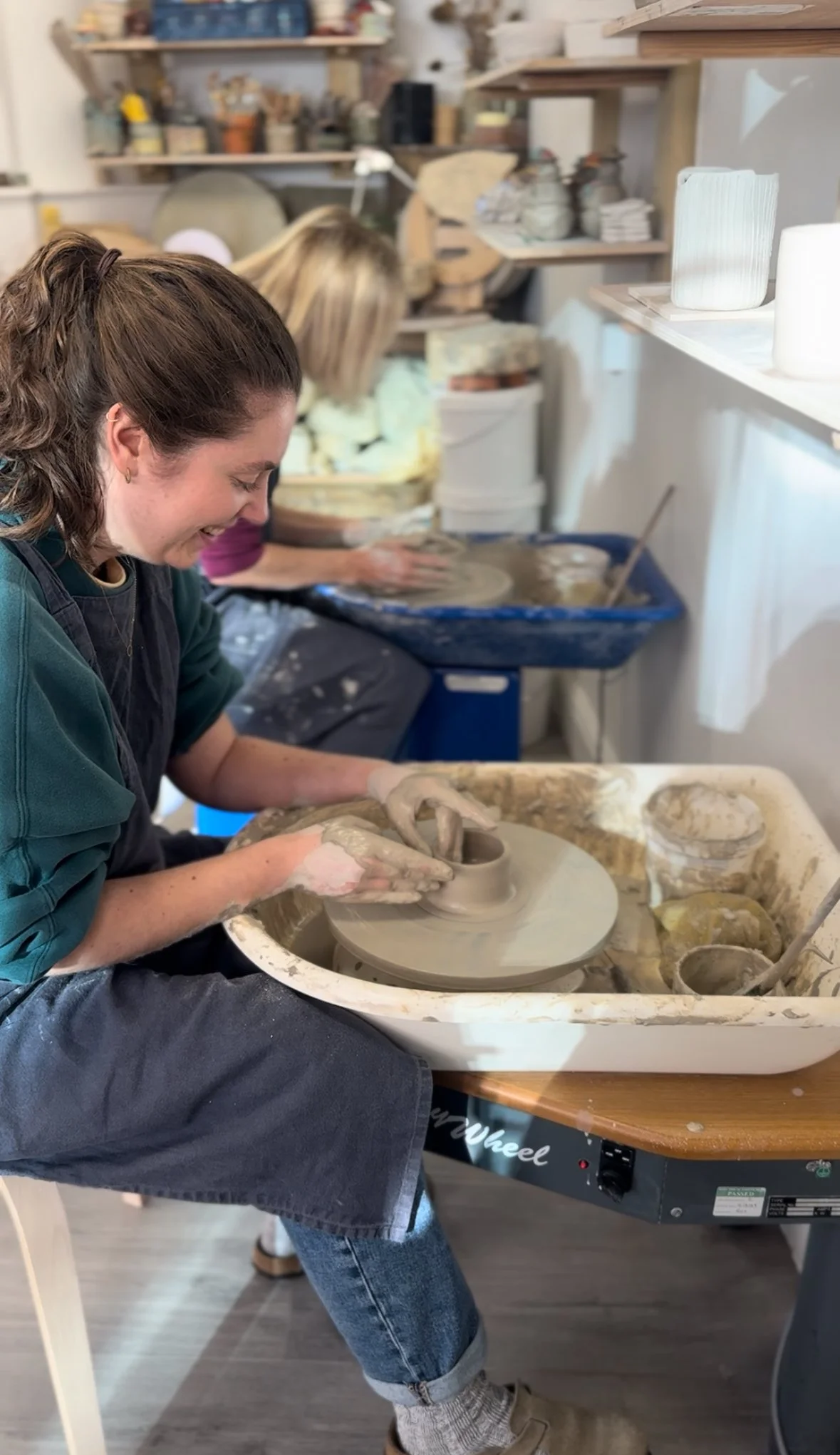 Two people laughing on pottery wheels (One-Day Throwing Course) Two participants laughing and shaping clay on pottery wheels during a One-Day Throwing Course at Clod Pots.