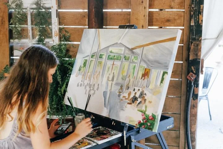 A woman painting an impressionistic scene of an indoor space with tall windows, plants, and people sitting at tables.