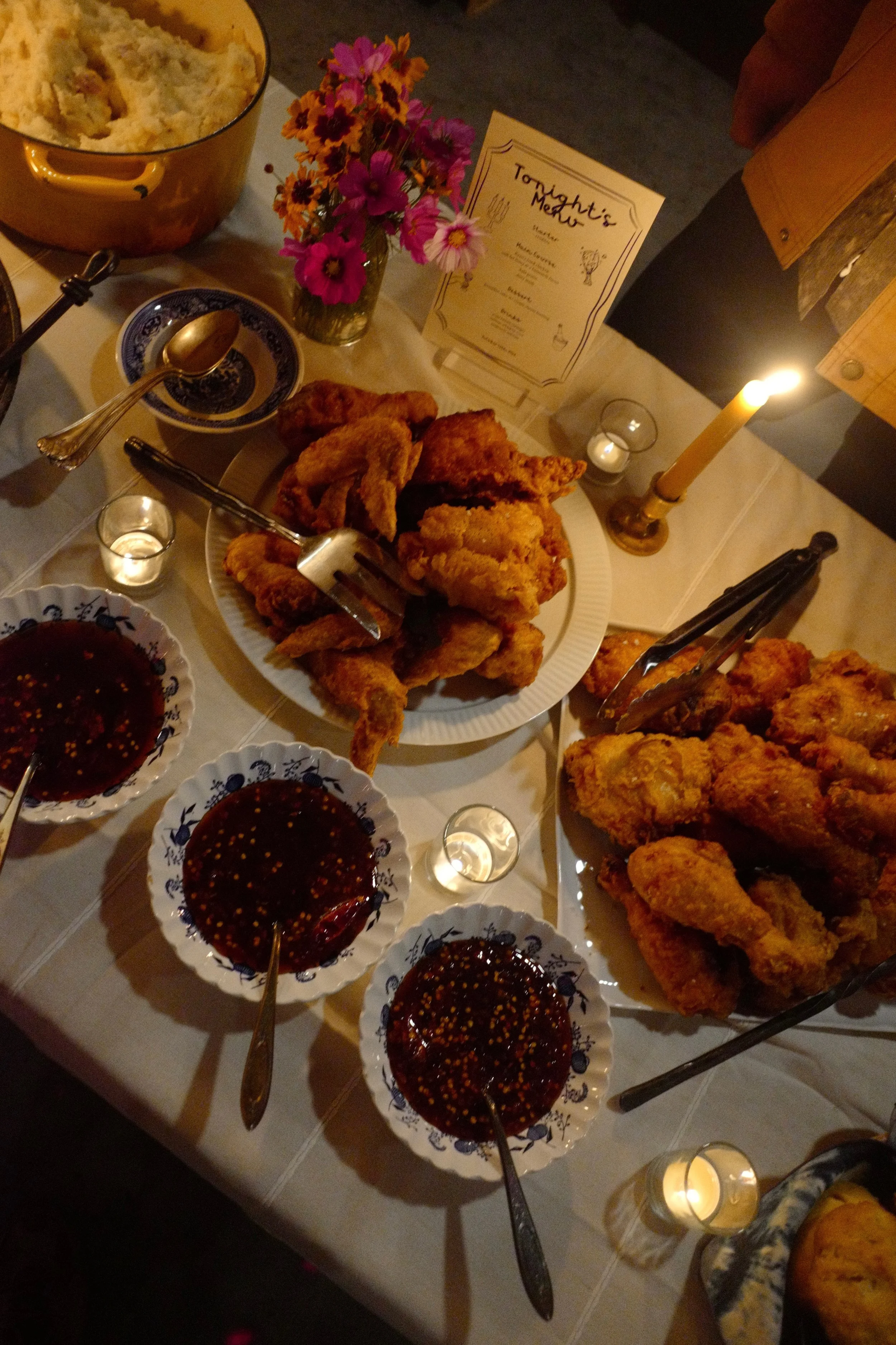 A table with fried chicken, bowls of dipping sauce, a bowl of mashed potatoes, a vase of flowers, and lit candles.